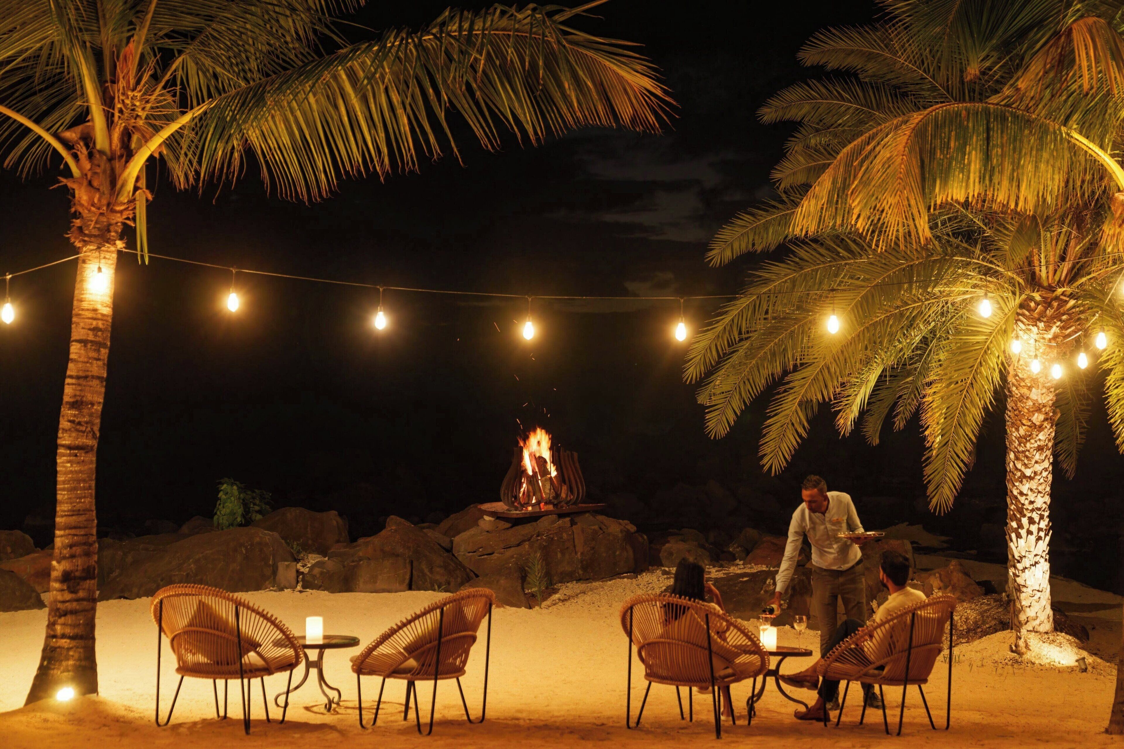 An outdoor dining area with string lights, palm trees, and a fire pit.