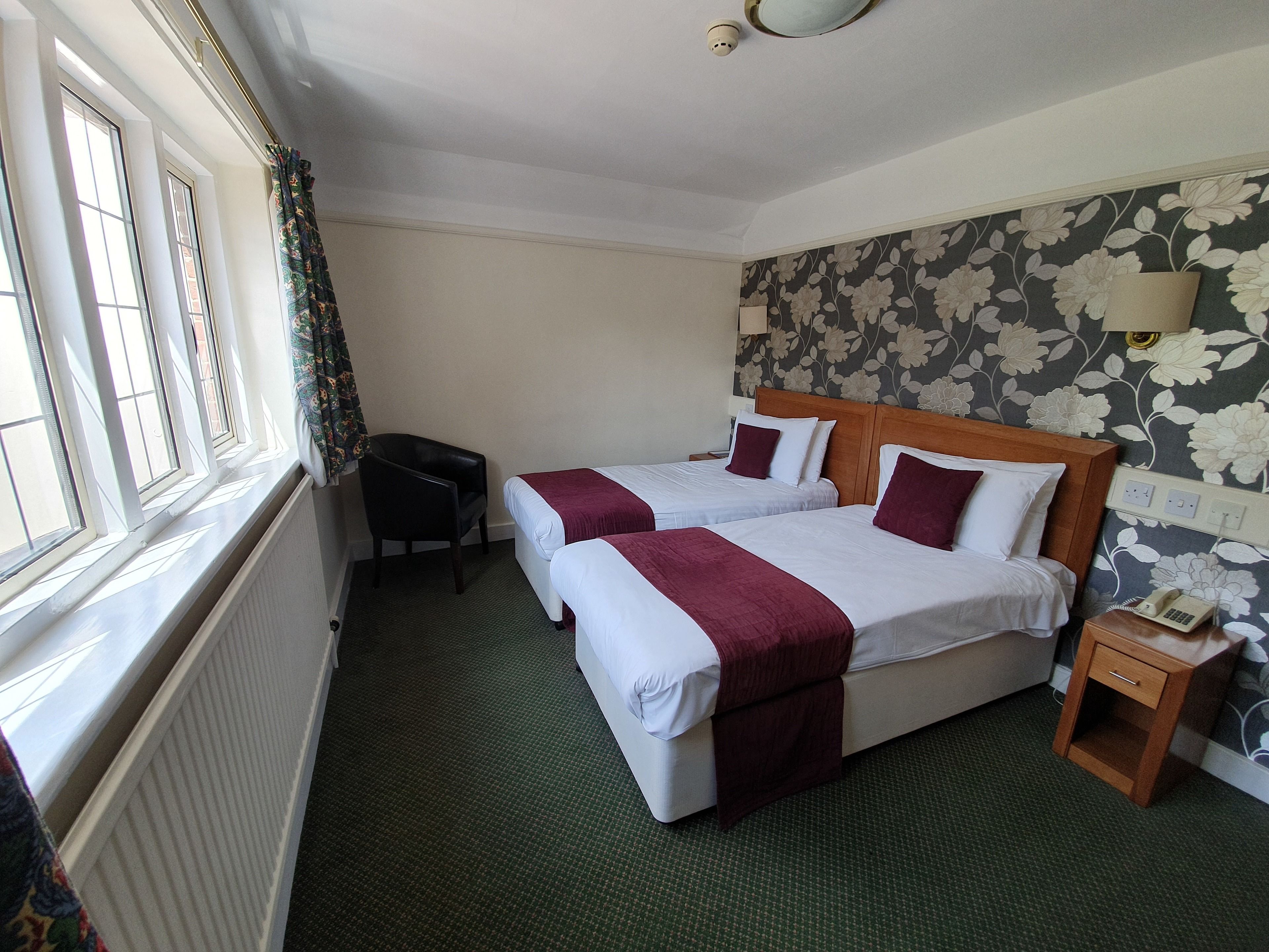A hotel room with two beds, a desk, a chair, a radiator, a telephone, and a window with curtains.