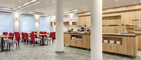 A modern hotel dining area with red and grey chairs, a central buffet station, and a large window with natural light.