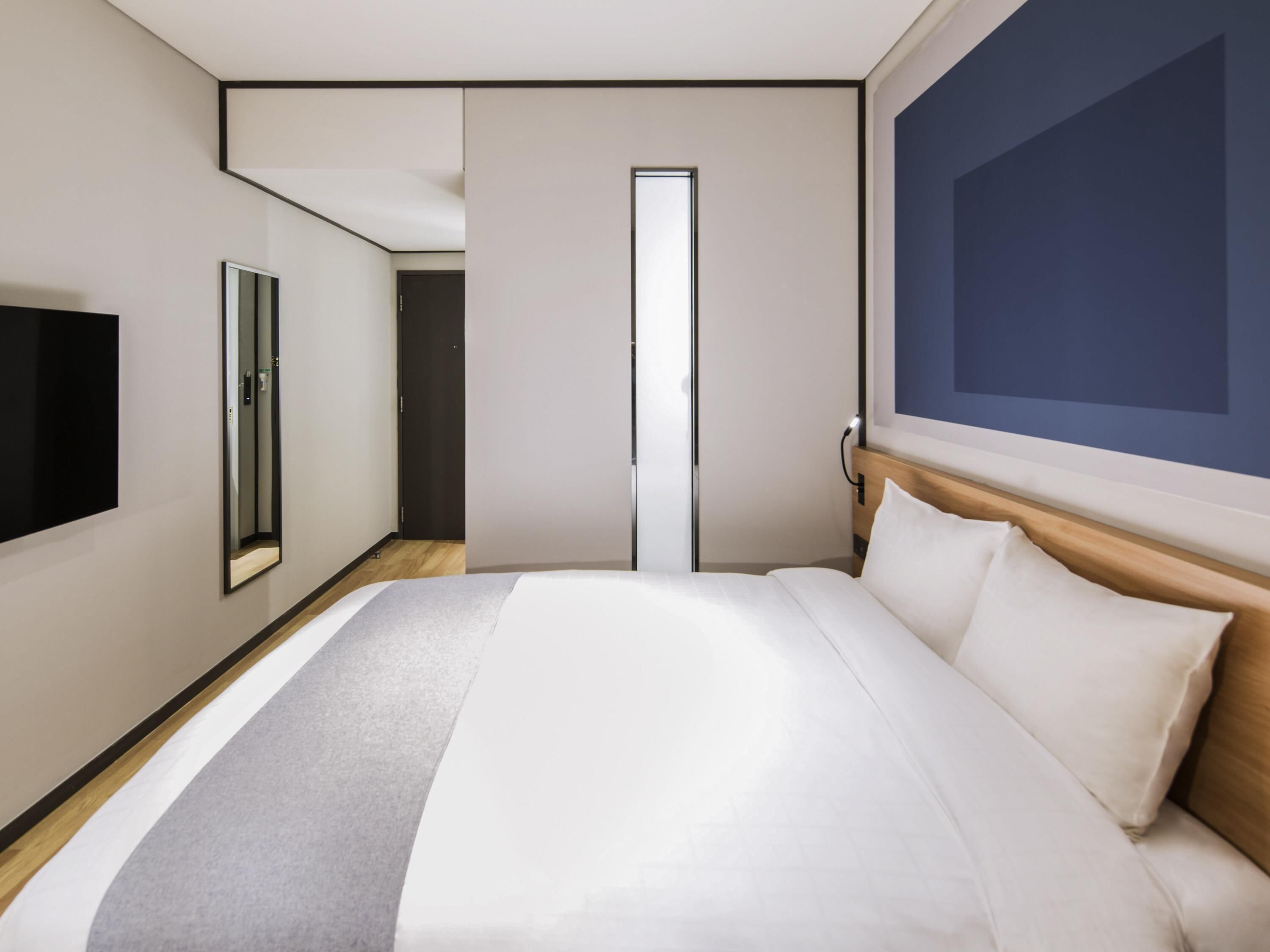 A modern hotel room with a large bed, a flat-screen TV, a mirror, and a large window with a view.
