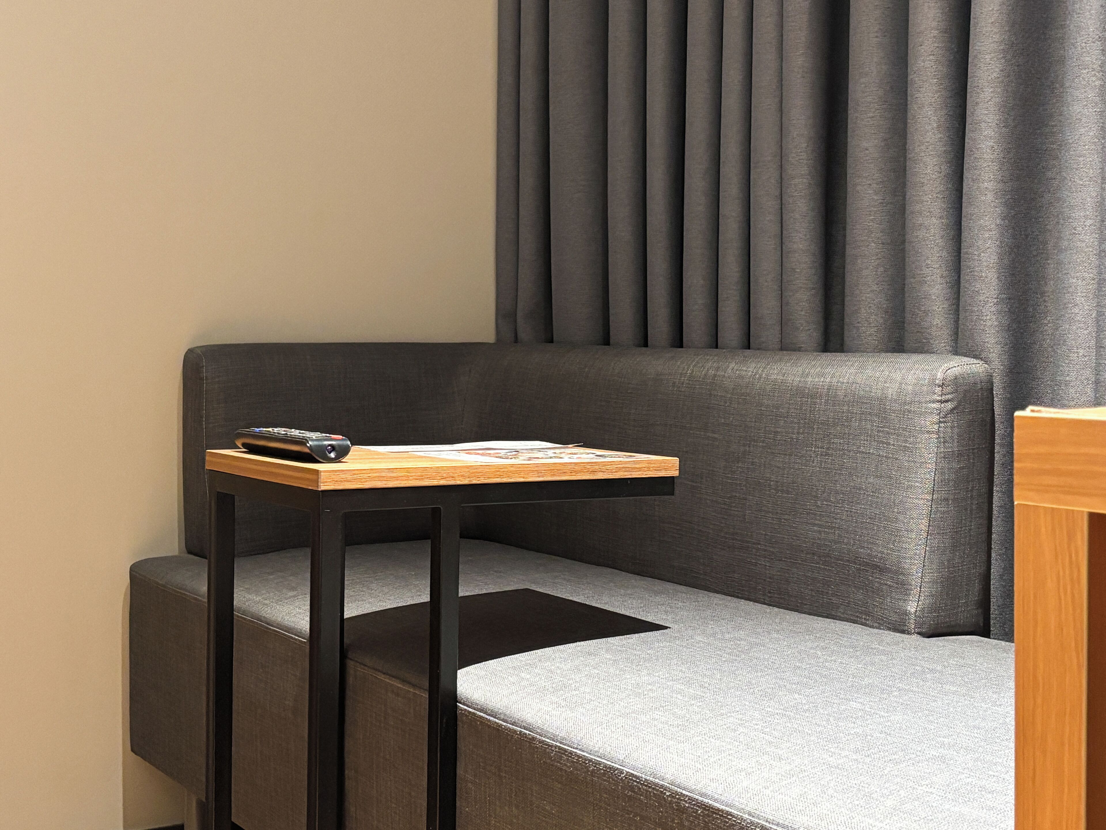 A modern hotel room with a grey sofa, a small wooden side table, and a remote control on the table.