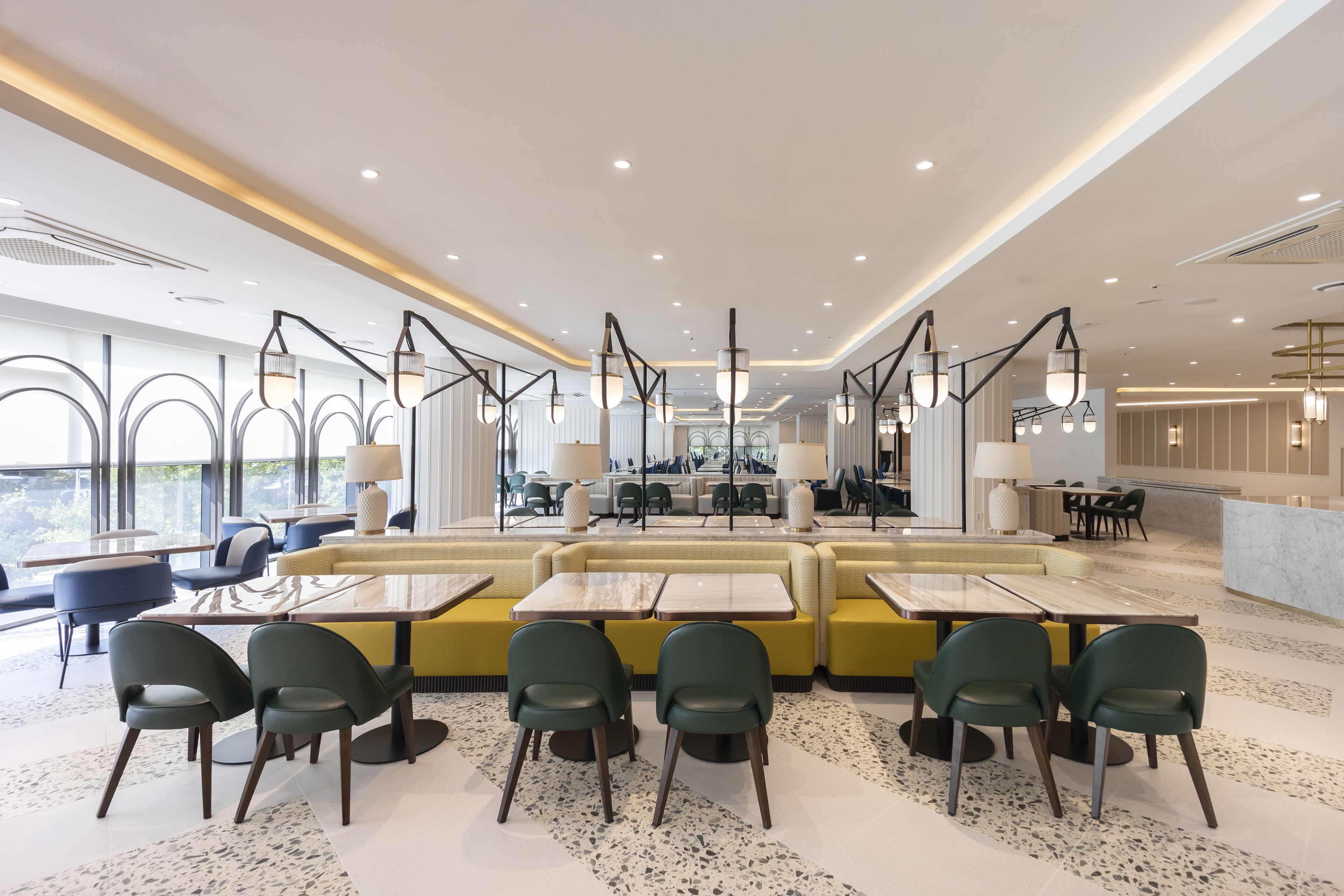 A modern restaurant with high tables and green chairs, a yellow seating area, and large windows offering a view.