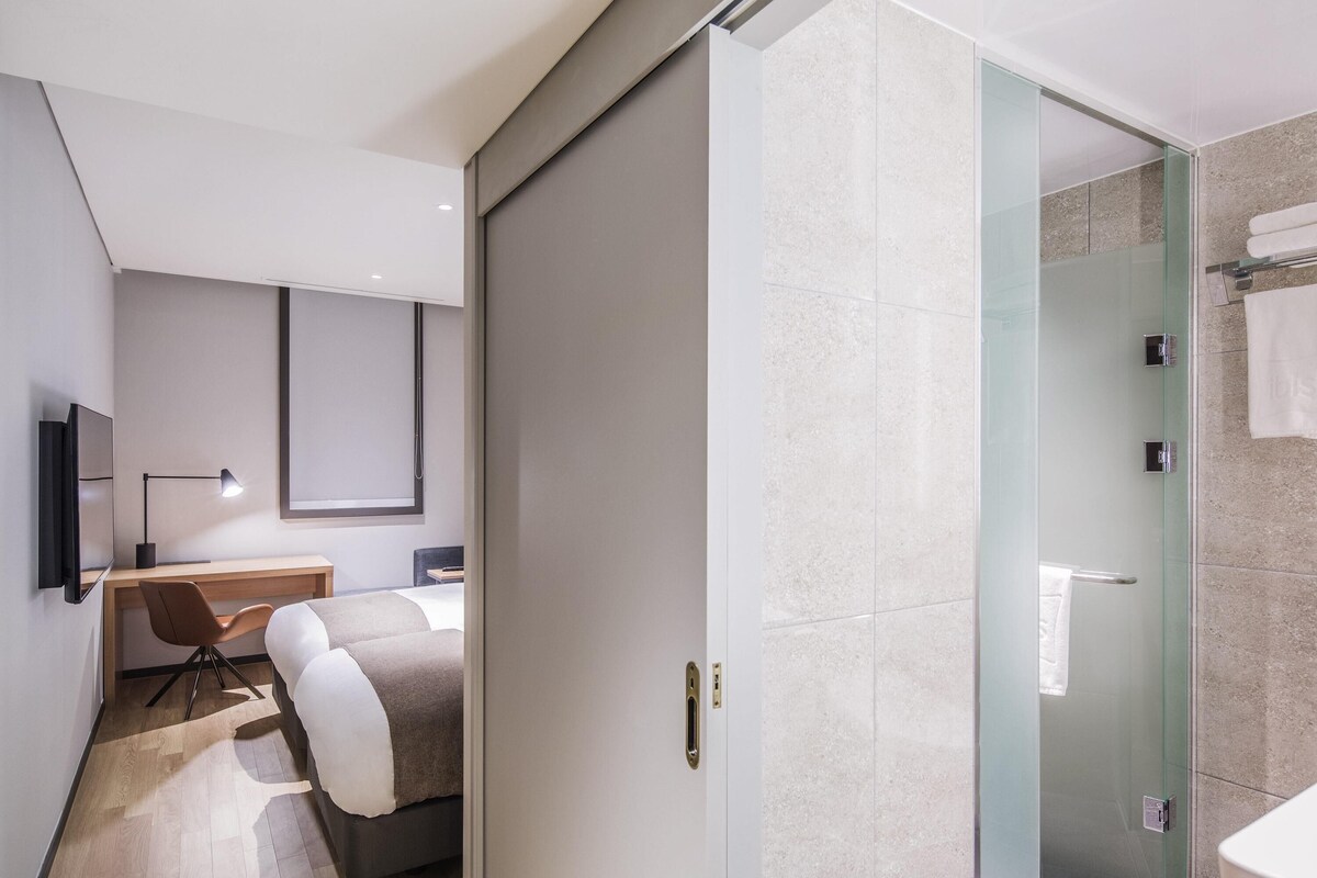 A modern hotel room with a bed, desk, chair, and a glass shower enclosure.