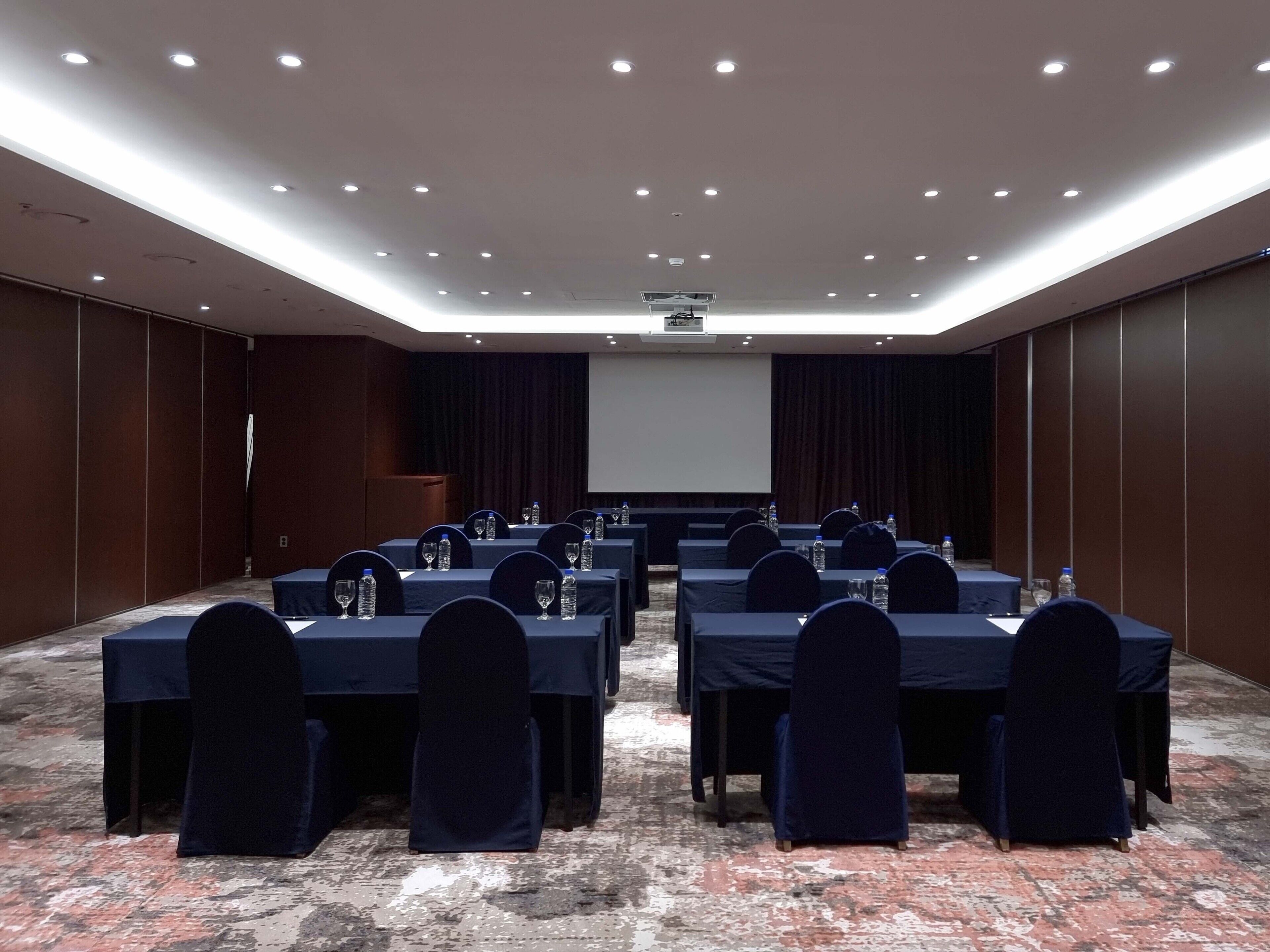 A conference room with tables set for a meeting, equipped with water bottles and glasses, and a projector screen at the front.