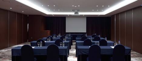 A conference room with tables set for a meeting, equipped with water bottles and glasses, and a projector screen at the front.
