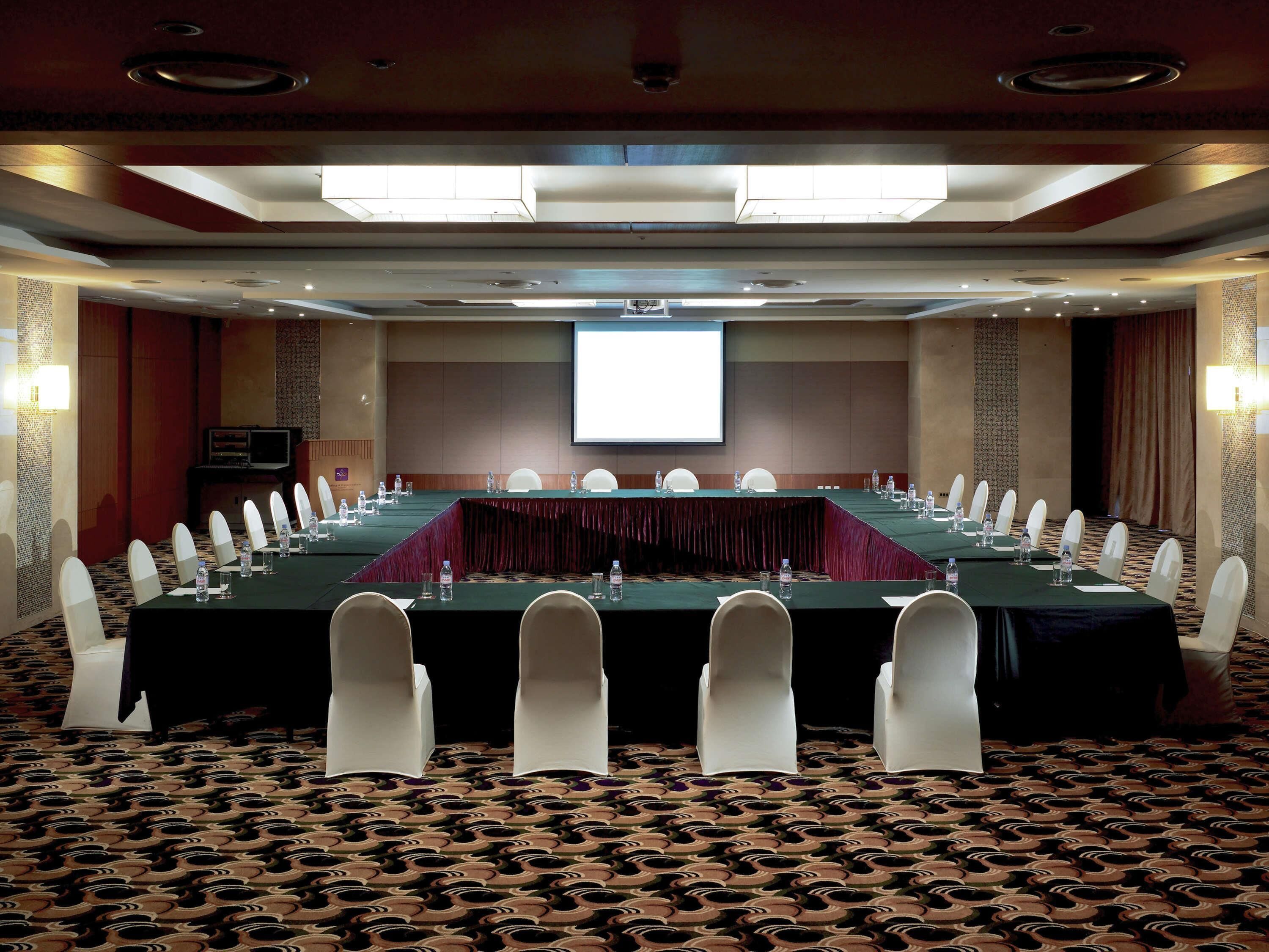 A conference room with a long table, white chairs, a projector screen, and a carpeted floor.