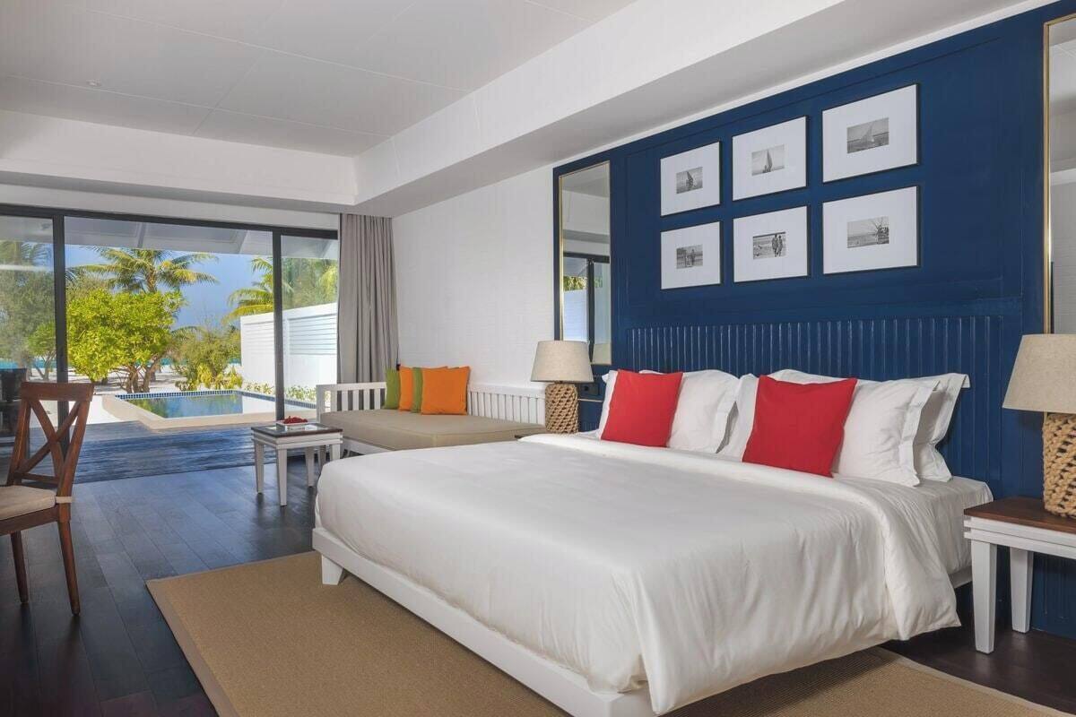 Una habitación de hotel moderna con una cama grande, un área de descanso con una silla y una mesa, vista a una piscina y una pared azul decorada con cuadros.