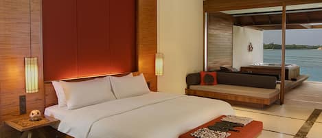 A modern bedroom with a large bed, a wooden nightstand, a bedside table with a lamp, a seating area with a sofa and cushions, and a view of the water.