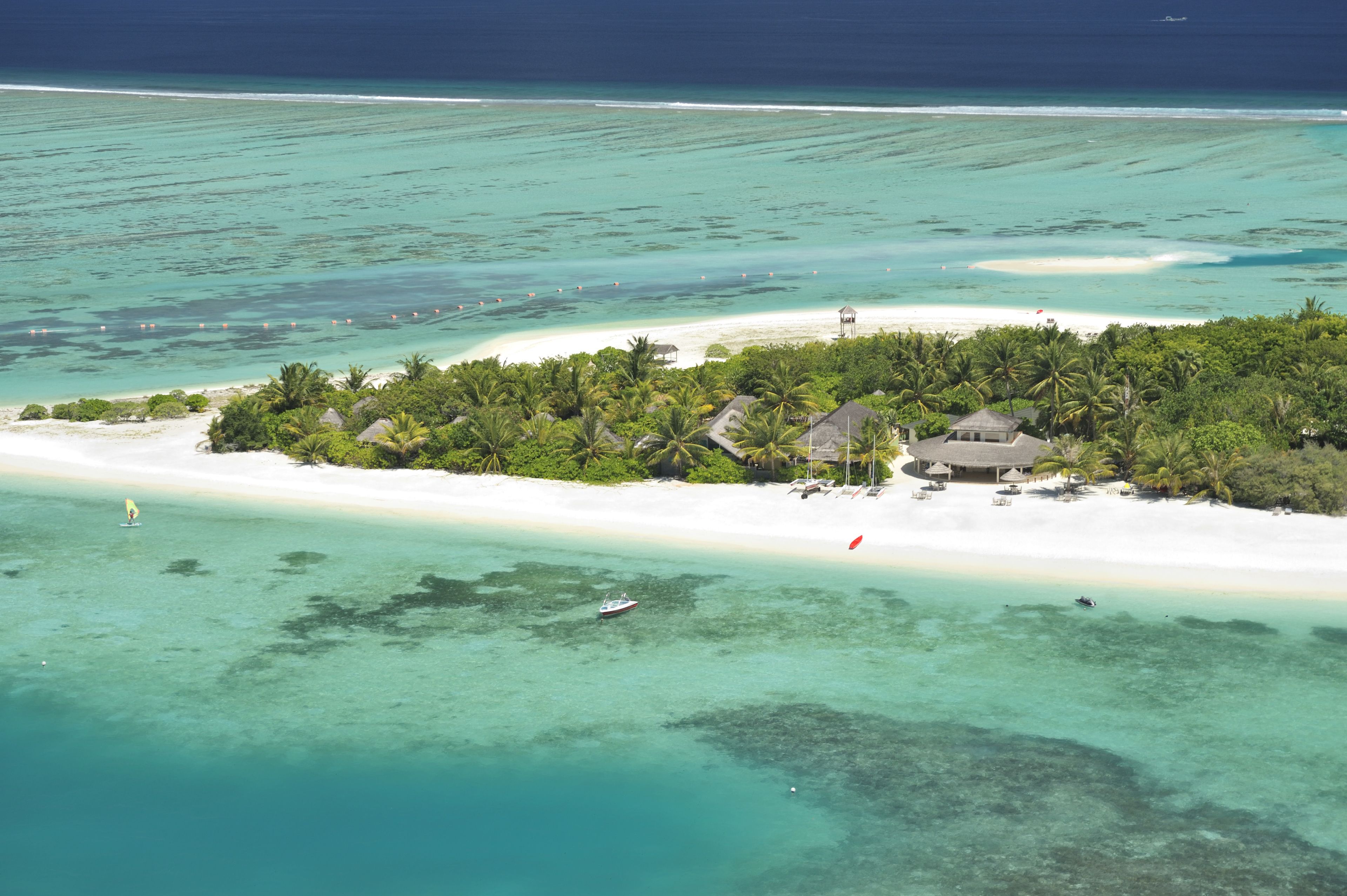 Vista aérea de una isla tropical con una playa de arena blanca, aguas turquesas cristalinas y un pequeño grupo de edificios rodeados de exuberante vegetación.