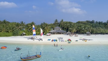 Ein Strand mit klarem türkisblauem Wasser, einem sandigen Ufer und Palmen. Boote und ein kleines Gebäude mit einem Strohdach sind zu sehen.
