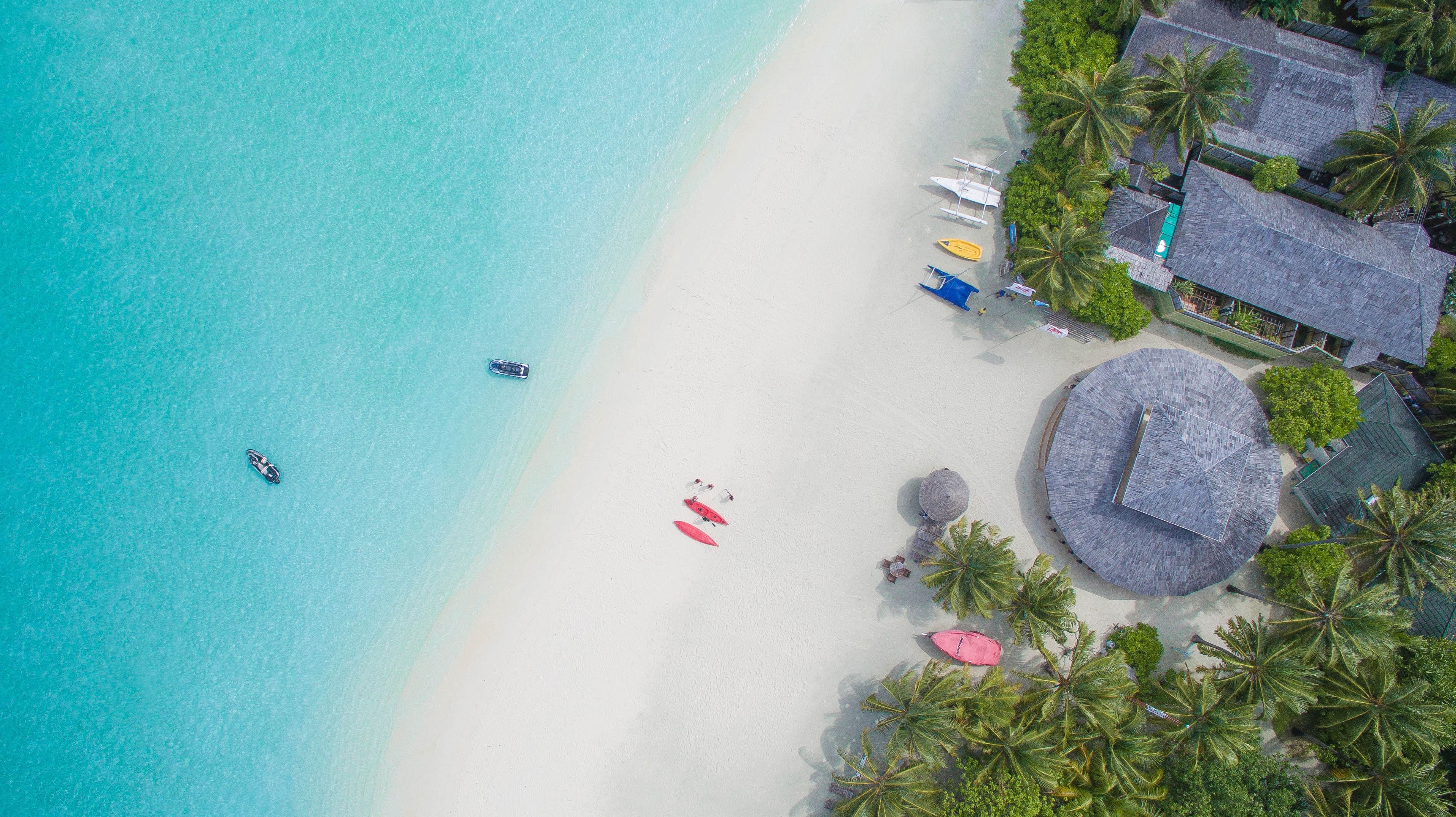 Vista aerea di una spiaggia con acque turchesi cristalline, una riva sabbiosa e diverse imbarcazioni. Si notano palme e un edificio con tetto di paglia nelle vicinanze.