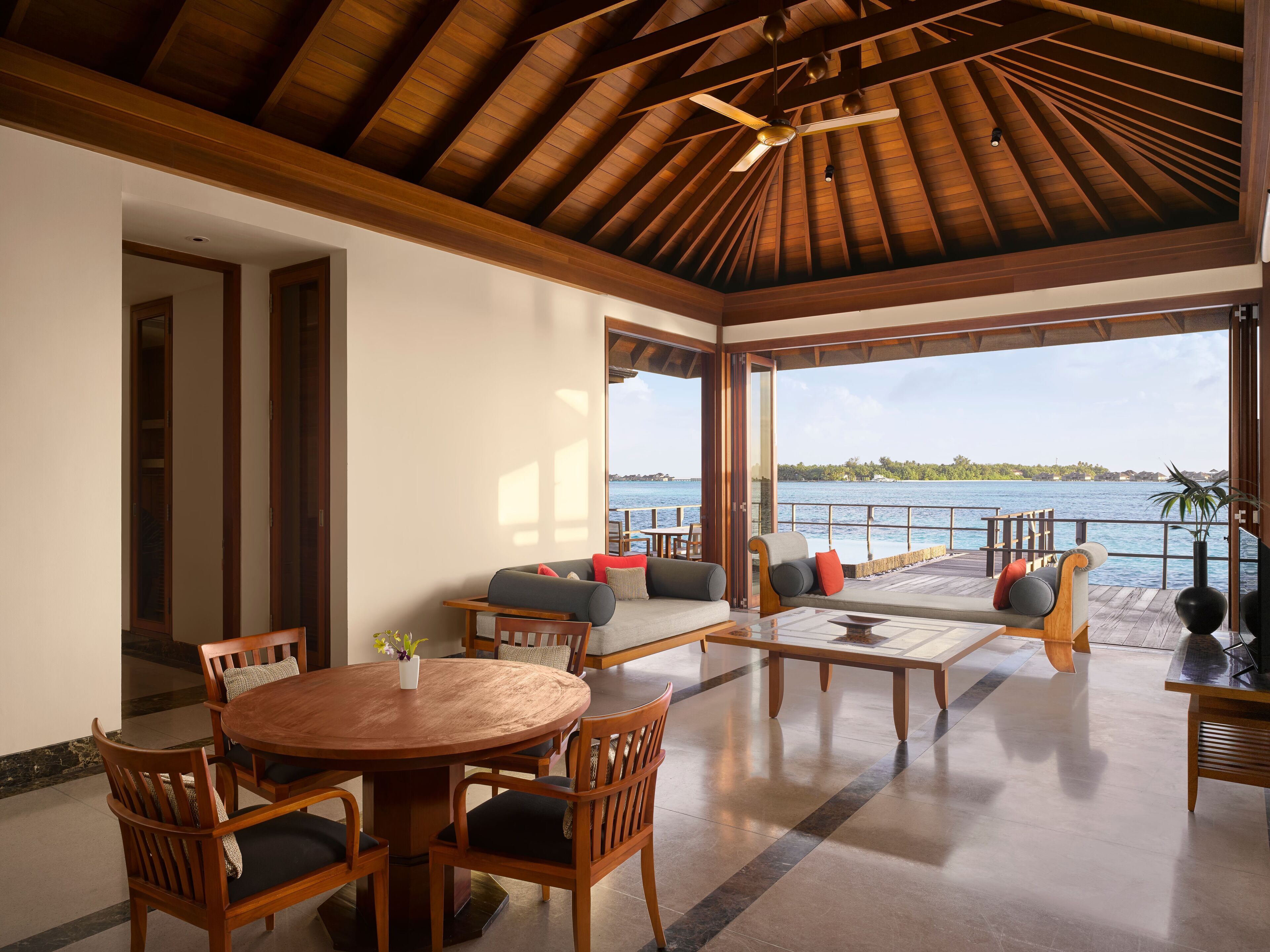 A spacious indoor area with a wooden ceiling, a round wooden table with chairs, a sofa with cushions, and a view of the sea through large windows.