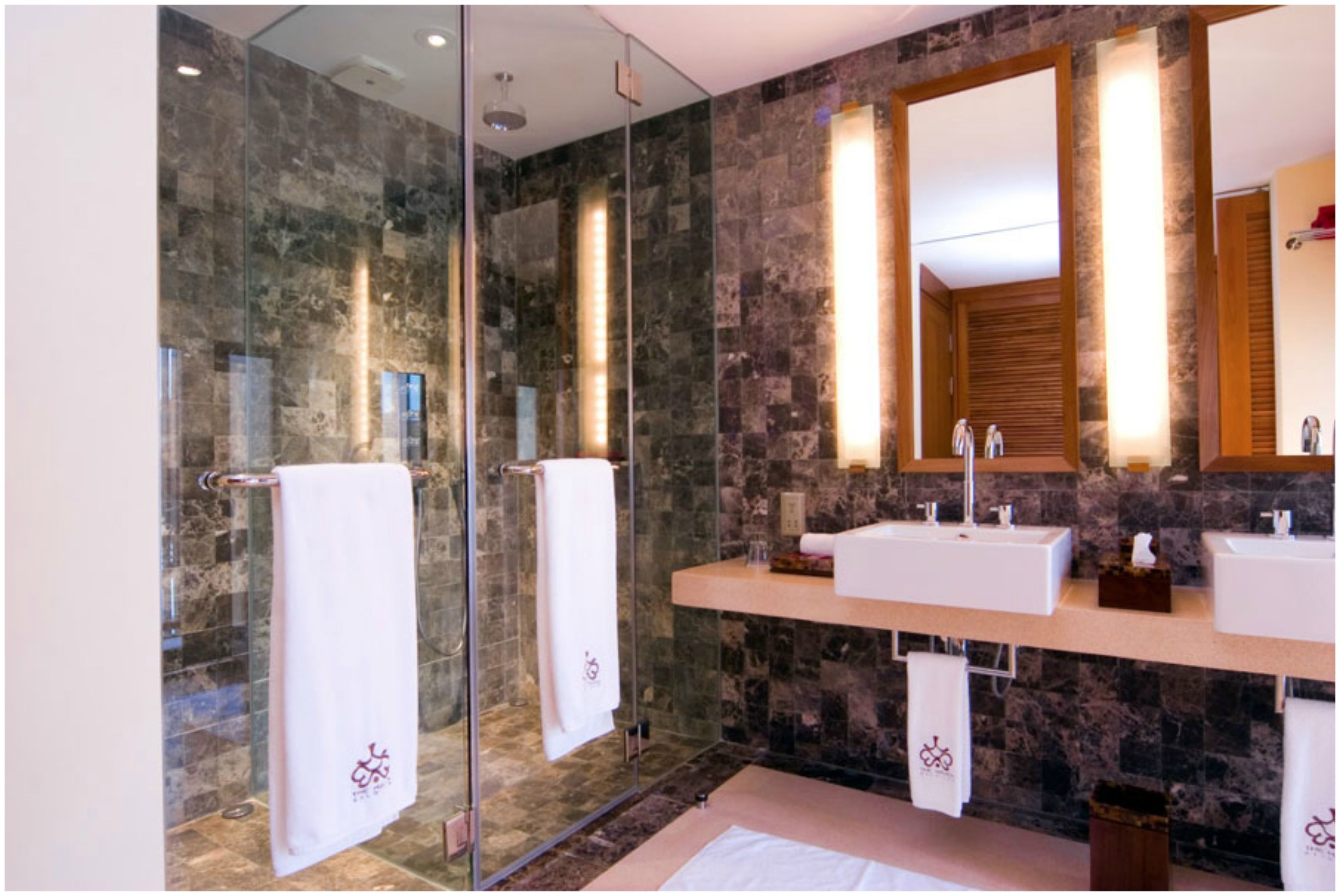 A modern bathroom with a glass shower enclosure, a marble wall, a wooden vanity with two sinks, and a large mirror.