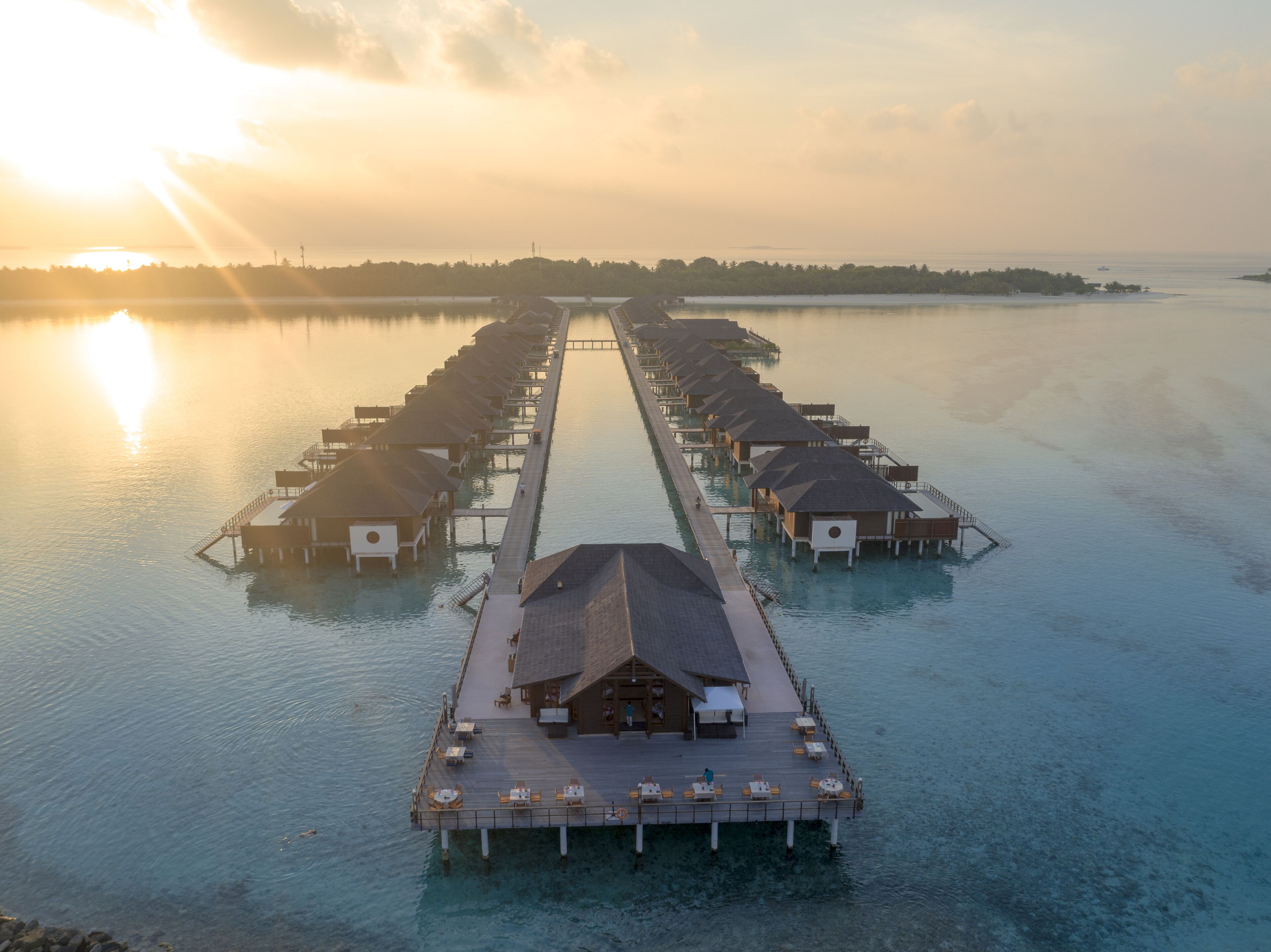 An aerial view of a row of overwater bungalows connected by a dock, with clear turquoise water and a sunset in the background.