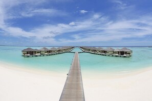 Eine lange Holzstege, der zu überwasser Bungalows führt, mit freiem Blick auf das türkisfarbene Meer und den weißen Strand.
