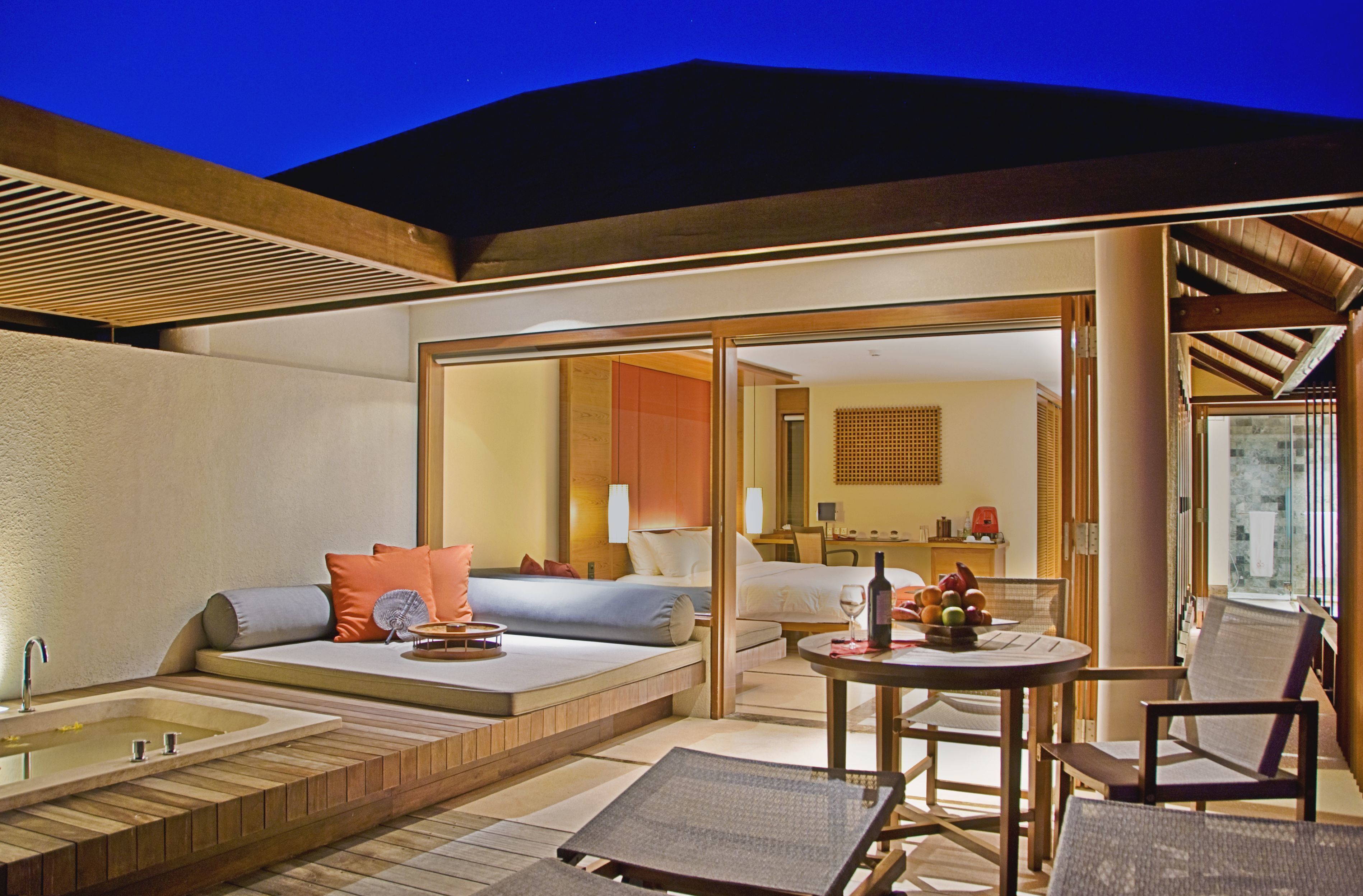 A modern hotel room with a large bed, a sitting area with a sofa and chairs, a dining table, and a large window offering a view of the outdoors.