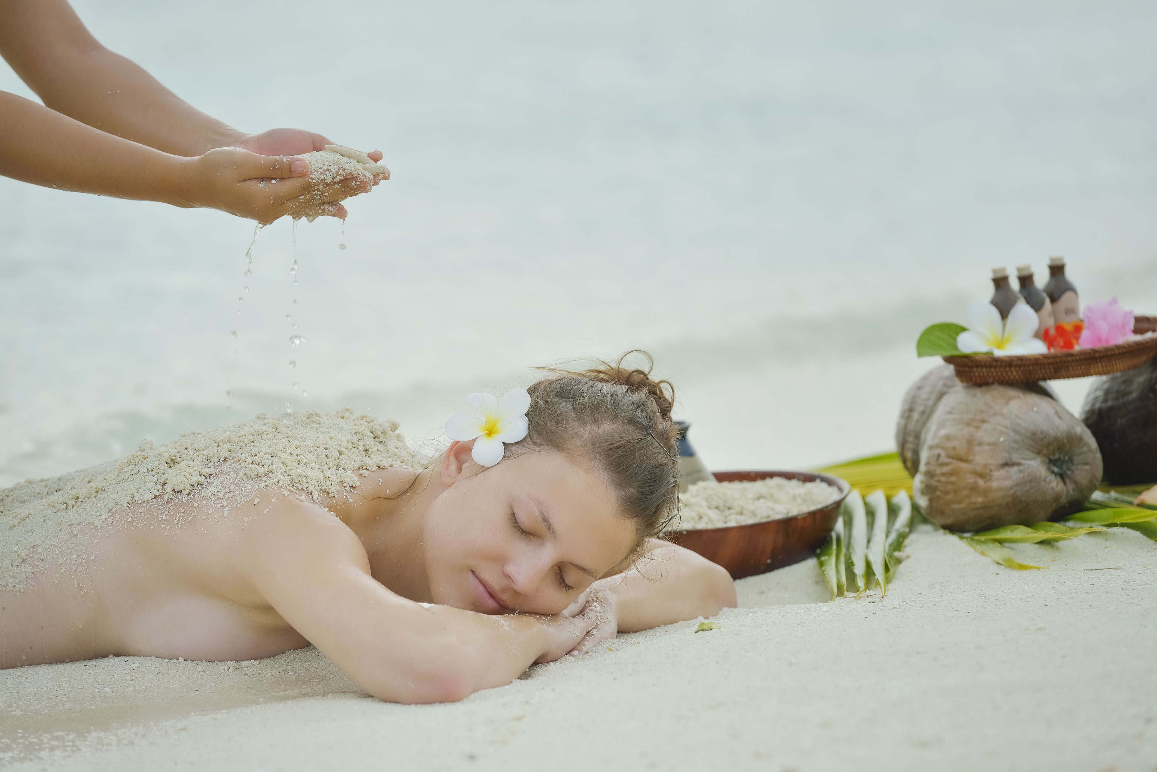 A person lying on a sandy surface with a flower in their hair, surrounded by a bowl of coconuts and bottles.
