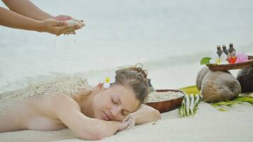 A person lying on a sandy surface with a flower in their hair, surrounded by a bowl of coconuts and bottles.