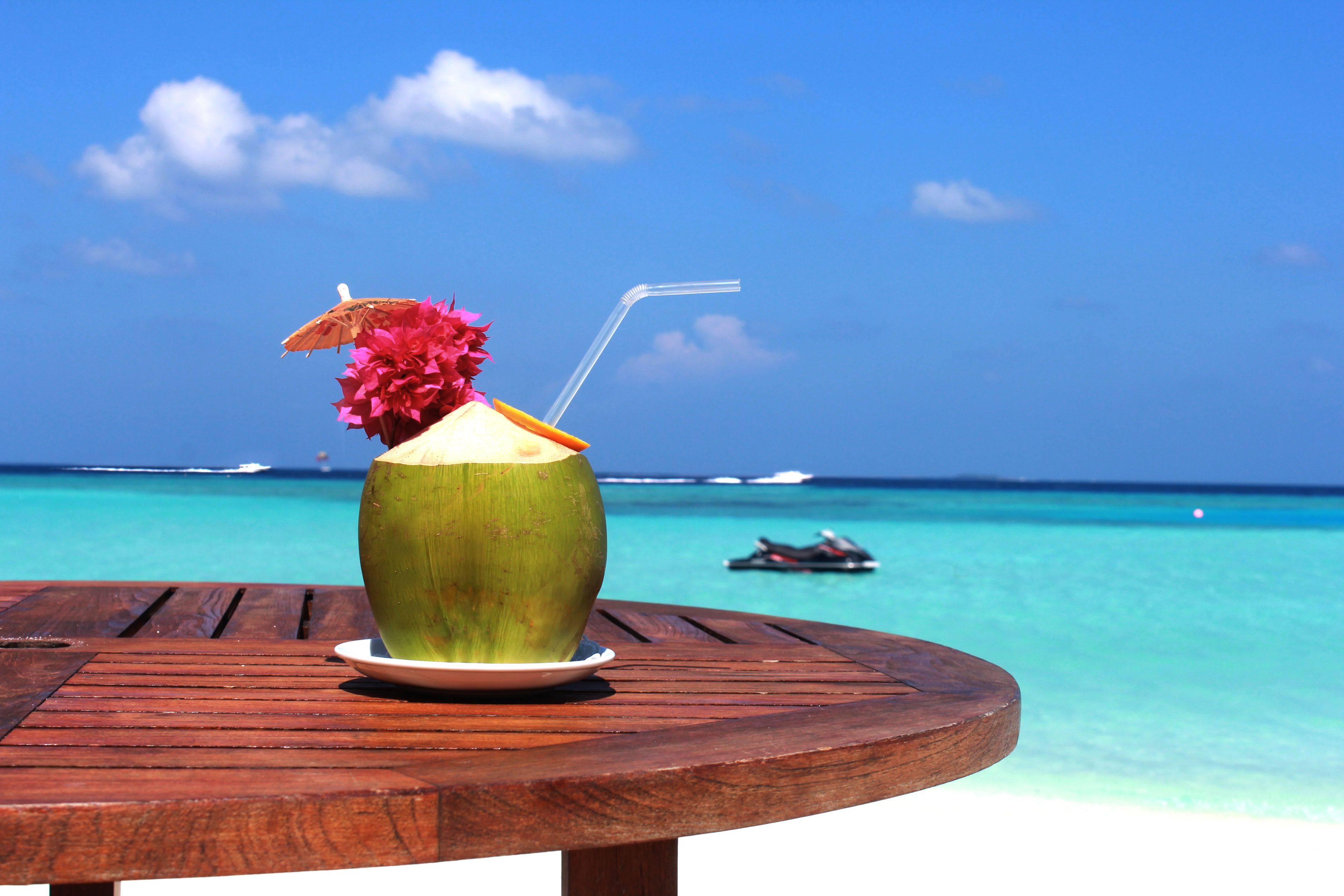 A wooden table with a coconut drink garnished with a flower and straw, set against a beach backdrop with clear turquoise water and a distant boat.