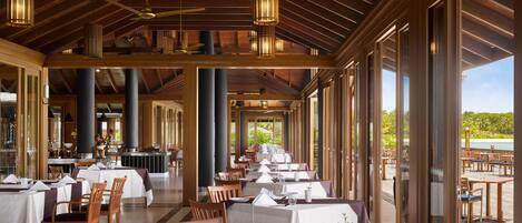 A spacious dining area with multiple tables and chairs, large windows offering a view of the outdoors, and a high ceiling with exposed beams and hanging lights.