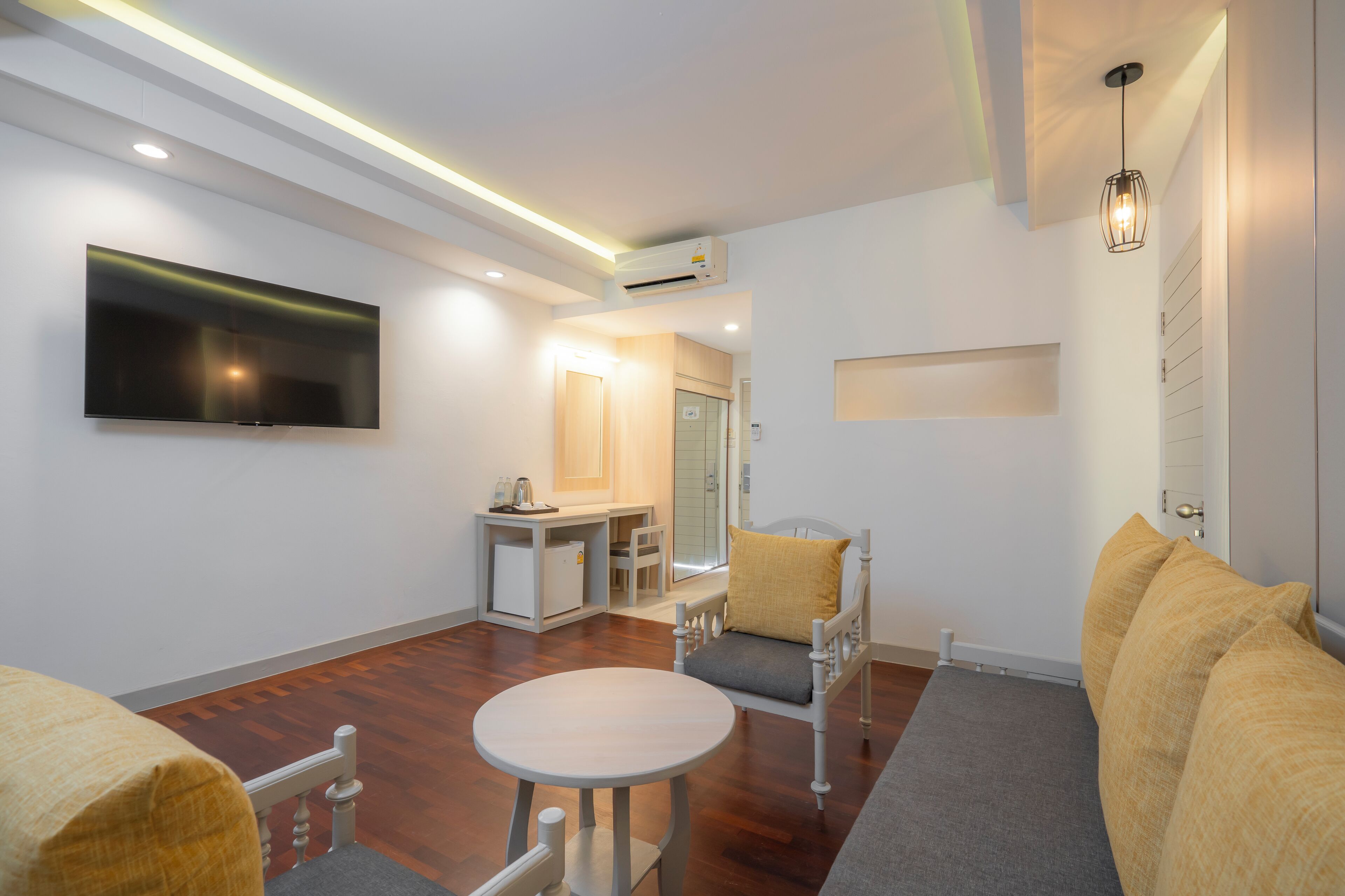 A modern hotel room with a flat-screen TV, a small kitchenette, a seating area with a sofa and armchairs, and a large window.