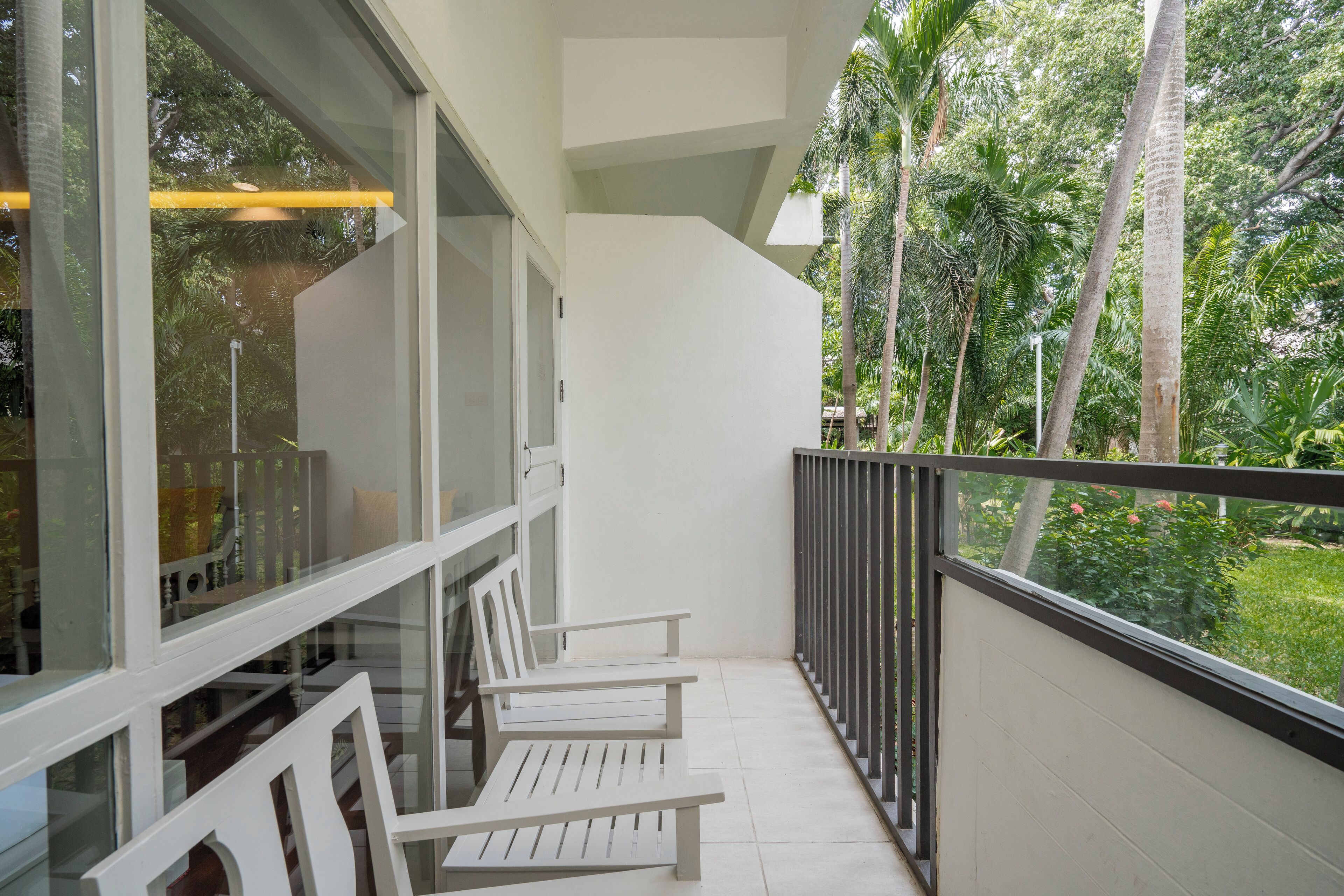 A balcony with white chairs and a glass railing overlooking a garden with palm trees.
