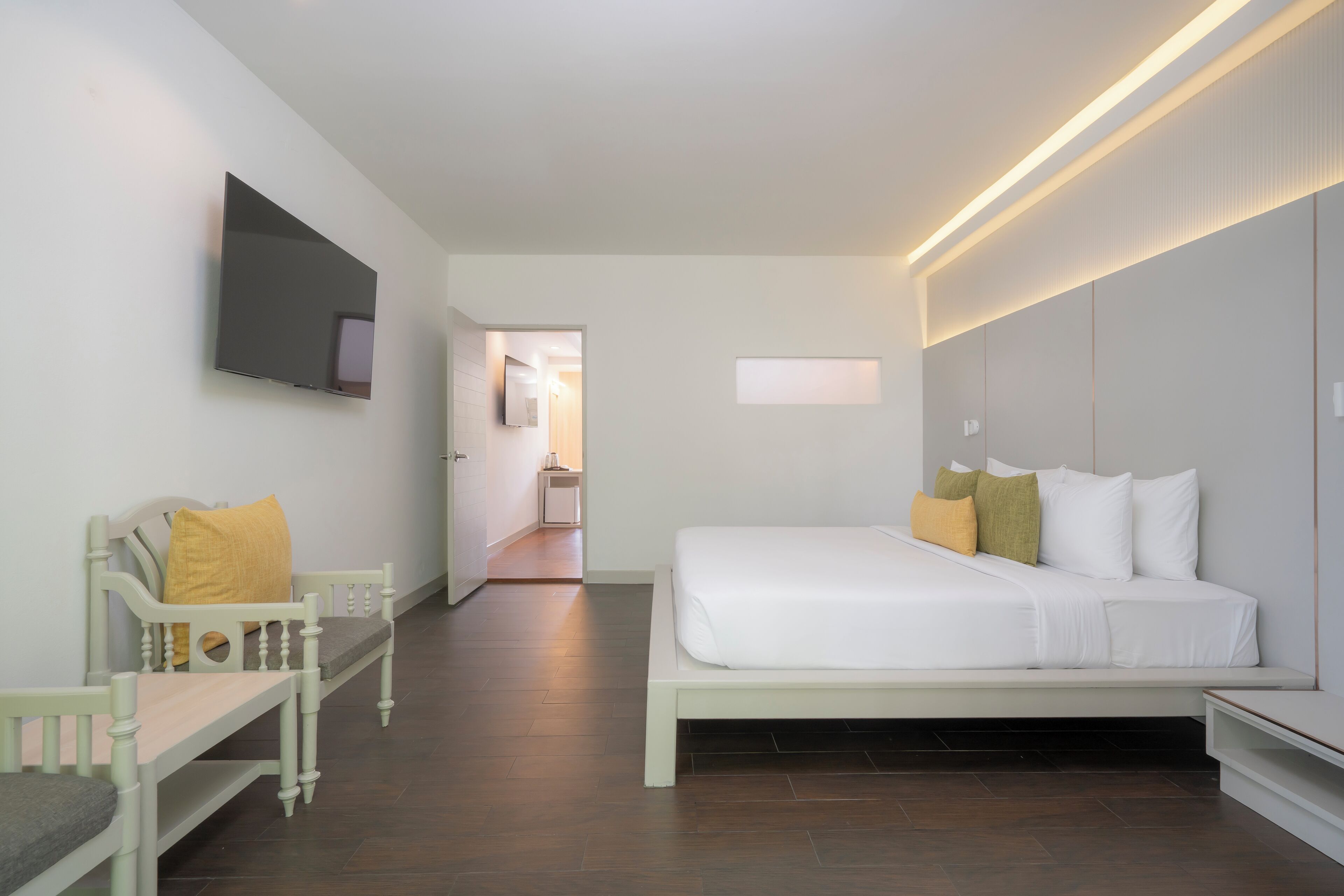 A modern hotel room with a large bed, a seating area with a bench, a flat-screen TV mounted on the wall, and a hallway leading to a bathroom.