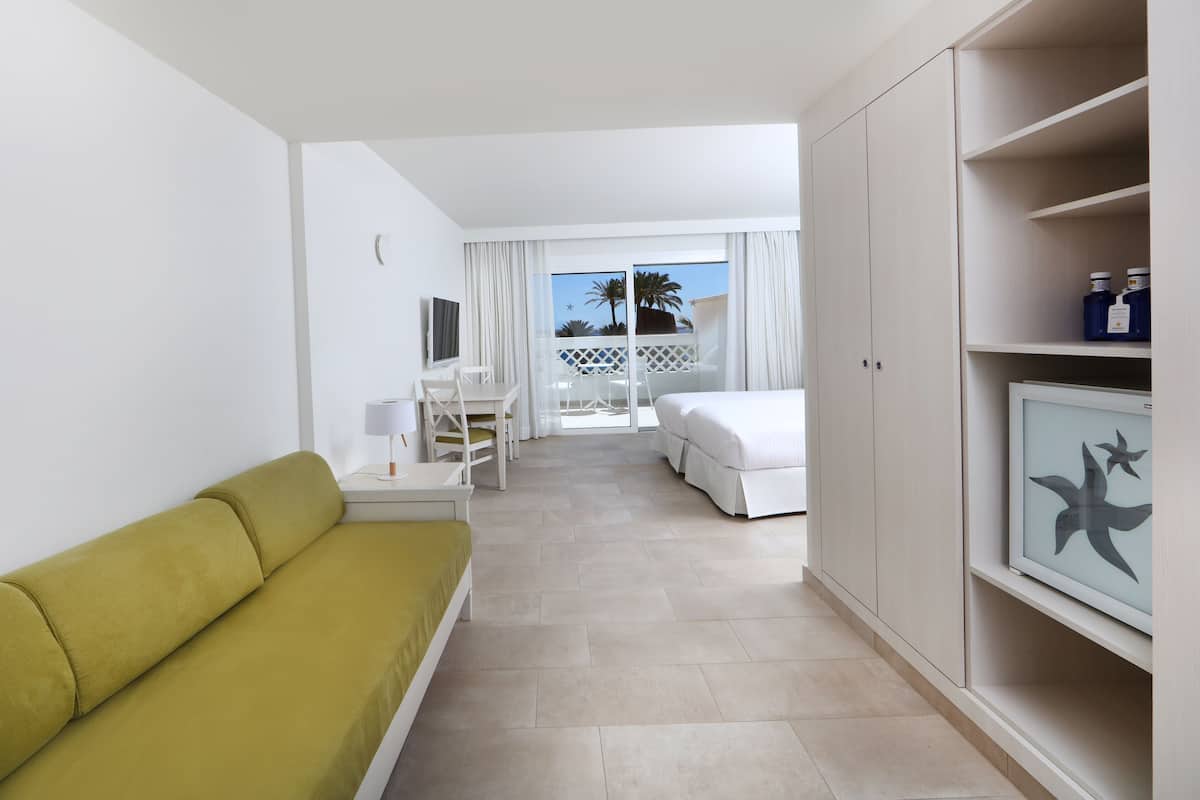 Habitación de hotel con sofá verde, cama blanca, escritorio y balcón con vistas a palmeras.