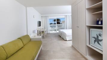 Une chambre d’hôtel avec un canapé vert, un lit blanc, un bureau et un balcon offrant une vue sur les palmiers.