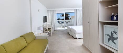 A hotel room with a green sofa, a white bed, a desk, and a balcony with a view of palm trees.