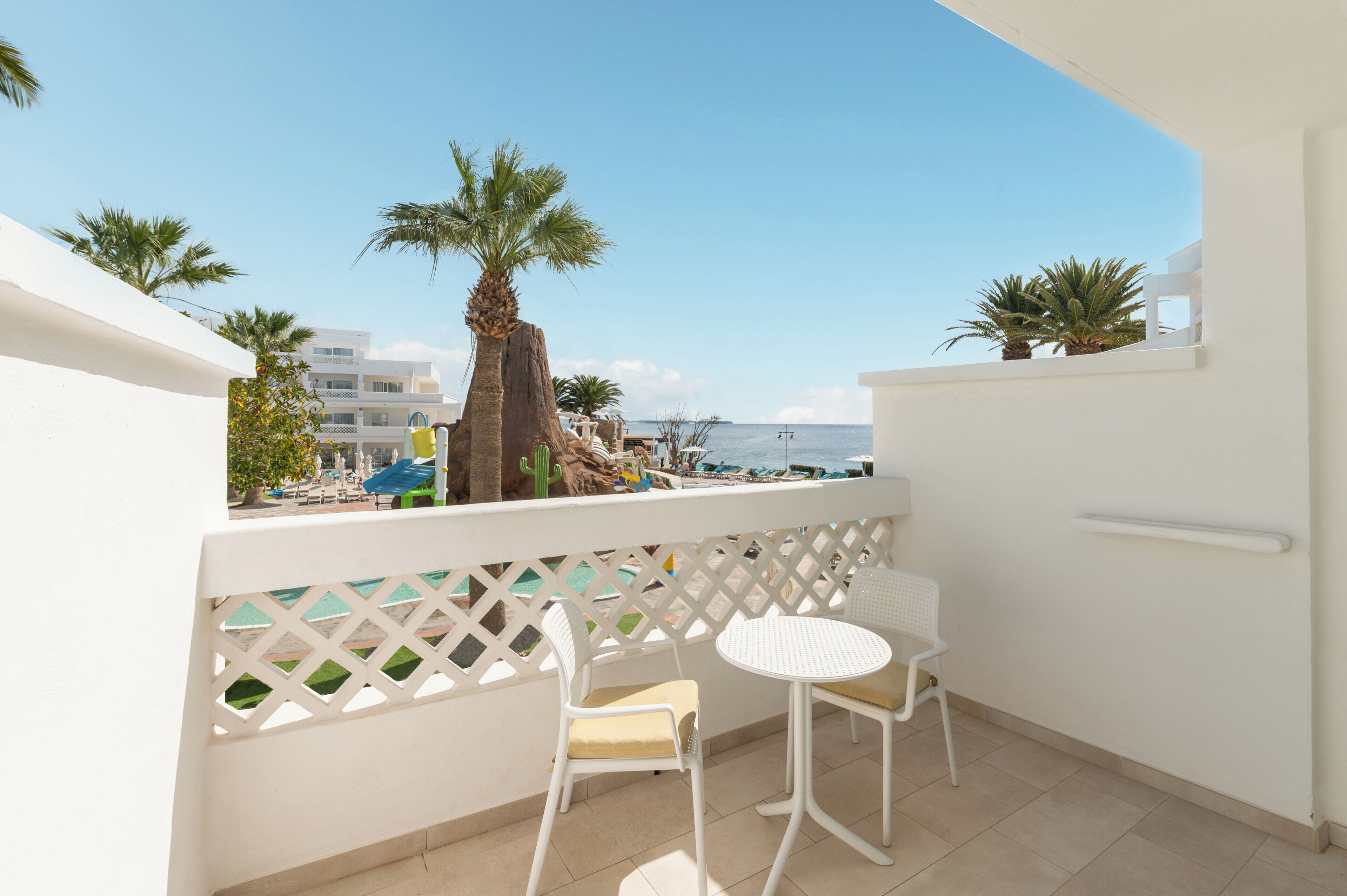 A balcony with two white chairs and a small table, overlooking a pool and palm trees.