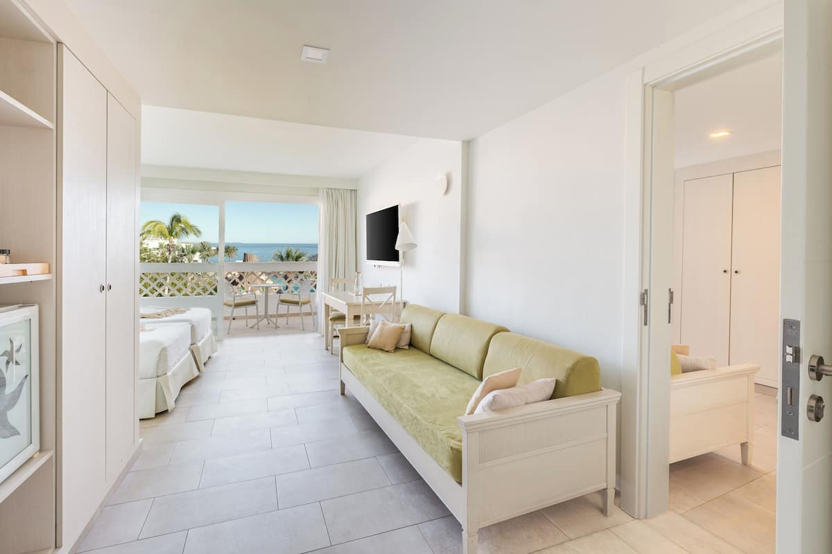 Habitación de hotel con cama, sofá, escritorio y televisor. Tiene un balcón con vista al mar.