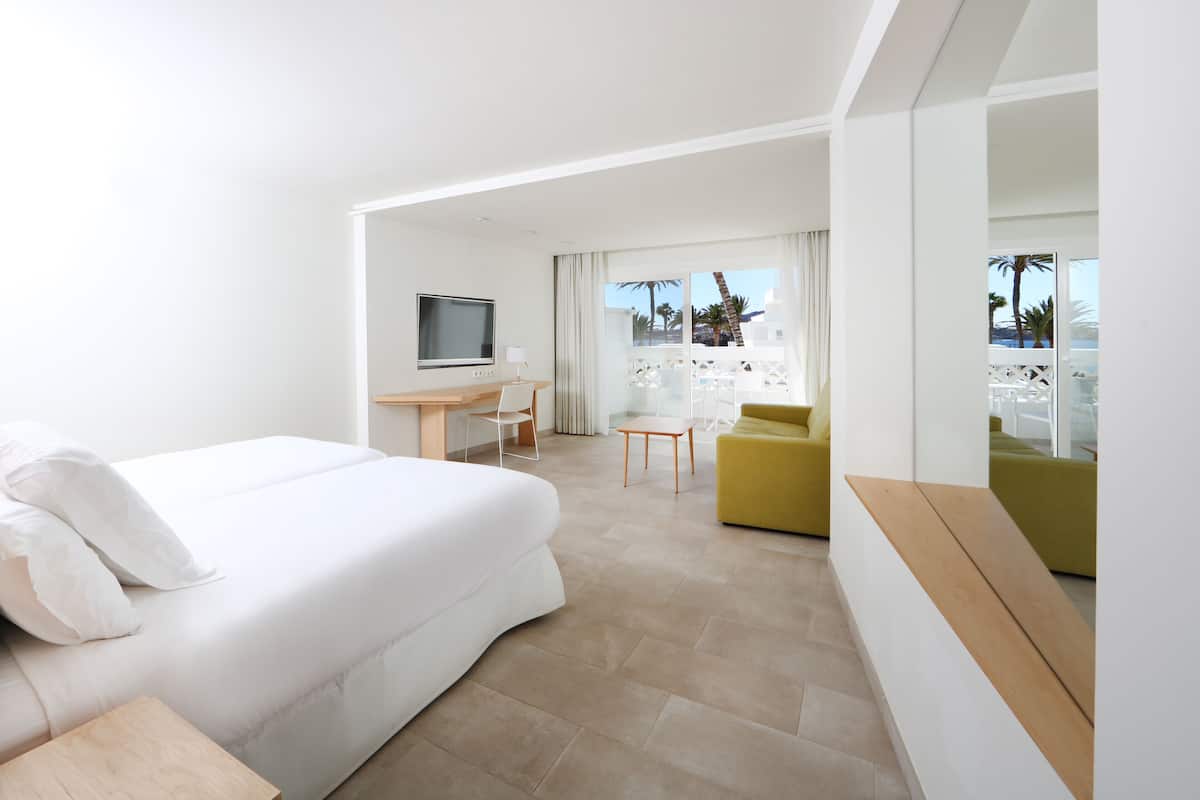 Una habitación de hotel moderna con una cama grande, un escritorio, un televisor, un sillón amarillo y un balcón con vistas a palmeras.