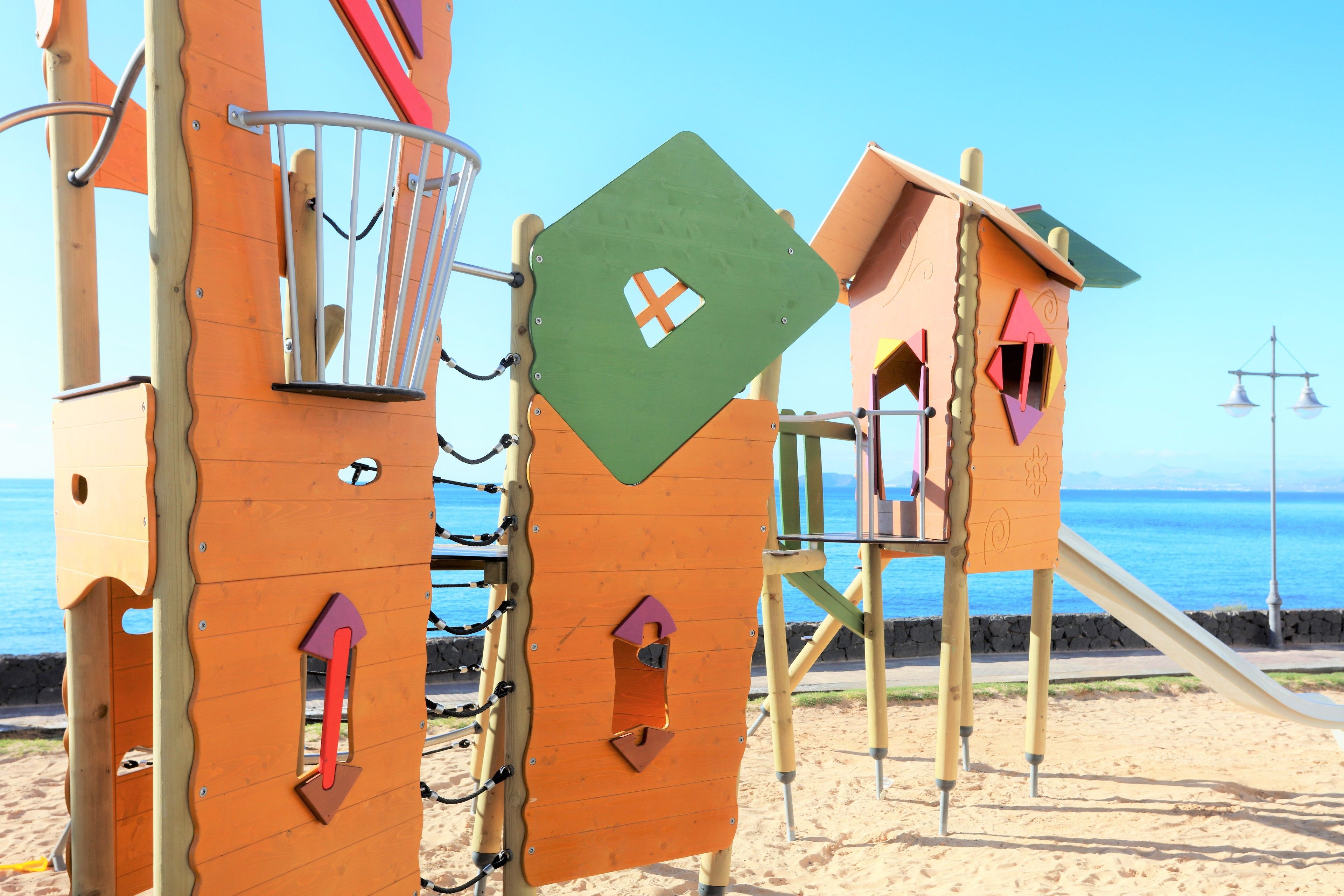 A children's play structure with climbing elements and a slide, located on a sandy beach with the ocean in the background.