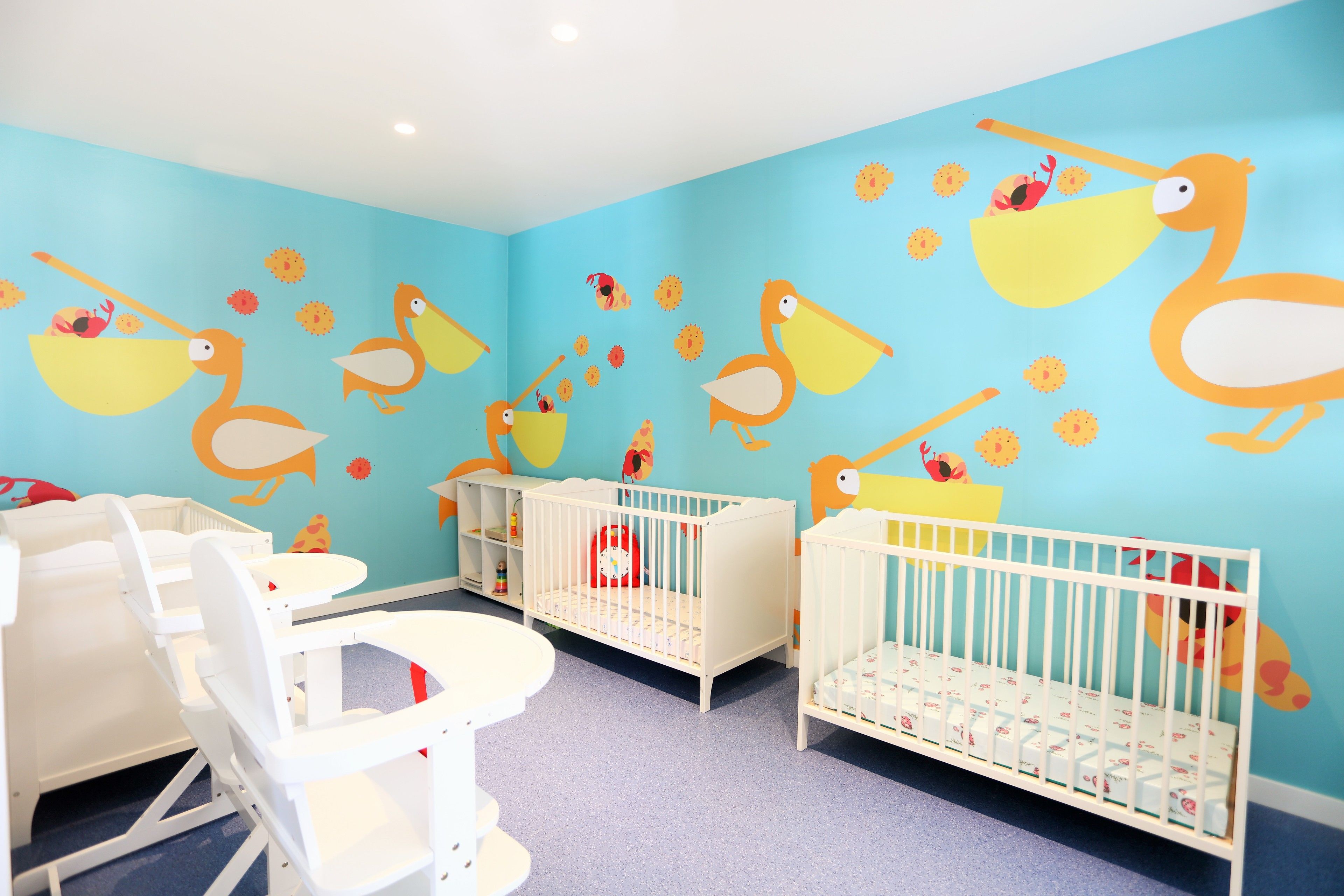 Une salle de jeux pour enfants avec des berceaux blancs, une petite table et un mur décoré de motifs d’oiseaux et de fleurs.