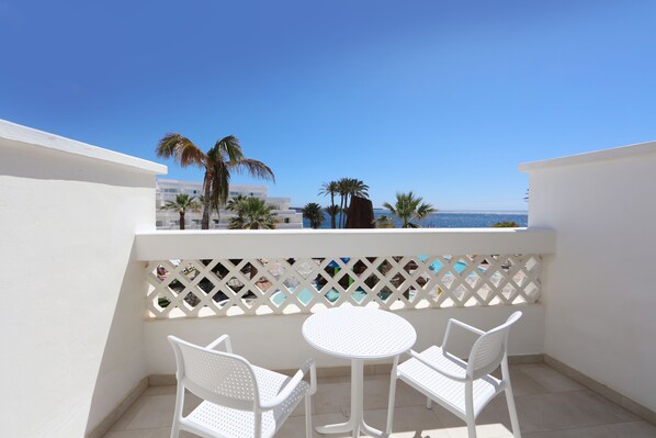Un balcon avec des chaises blanches et une table, d’où l’on peut admirer un ciel bleu dégagé et une vue lointaine sur des palmiers et des bâtiments.