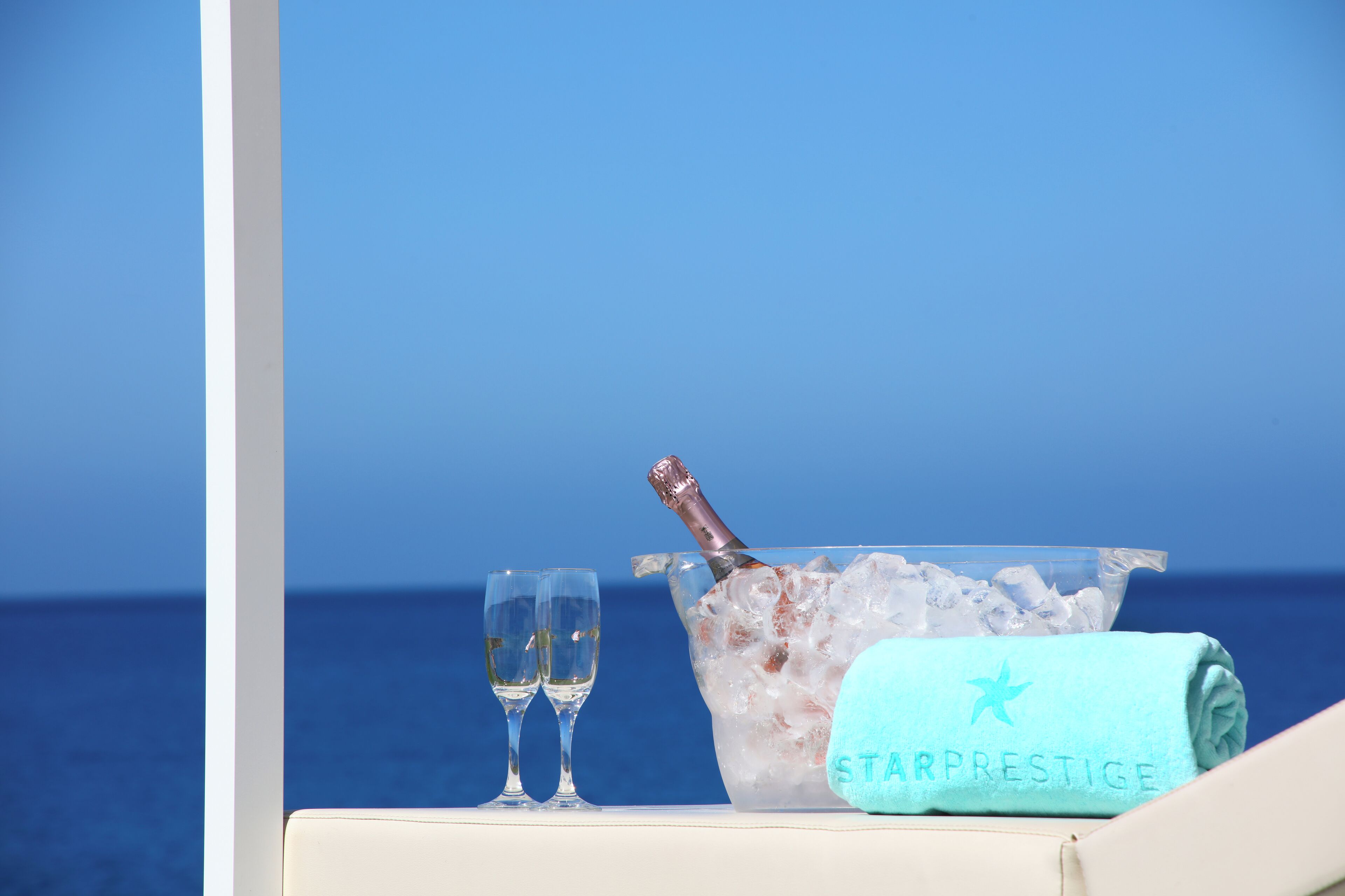 Una mesa en la playa con un balde de hielo, dos vasos y una toalla con la marca 'STARPRESTIGE'.
