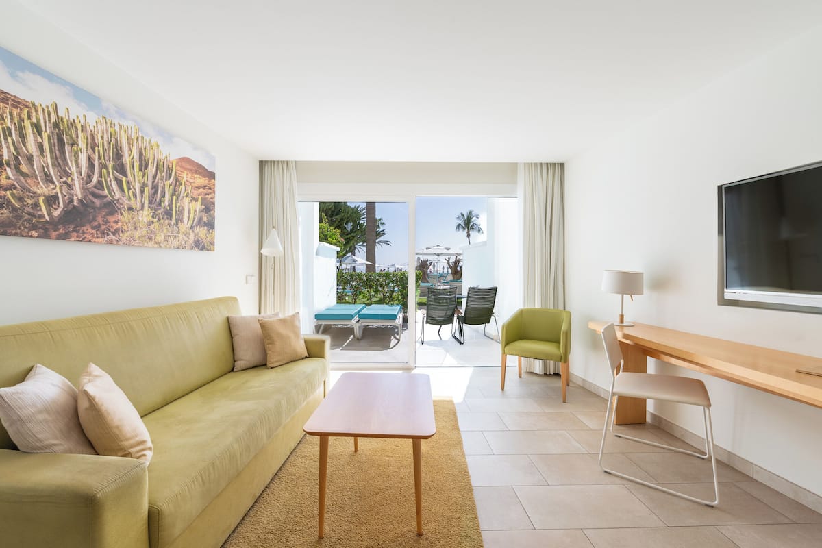 Una sala de estar con sofá, mesa de centro, televisor, escritorio y una silla. Hay un ventanal con vistas al exterior.