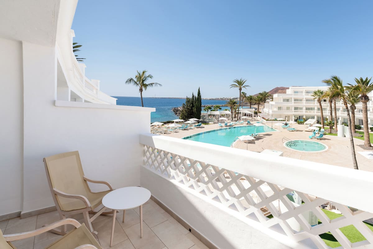 Un balcón con vista a la piscina, muebles blancos y cielo despejado.