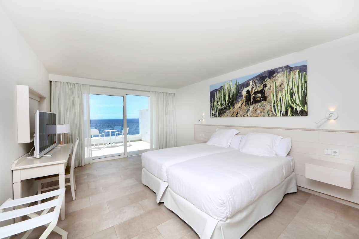Habitación de hotel con una cama, un escritorio con televisor, una silla, una ventana con cortinas y un cuadro de cactus en la pared.