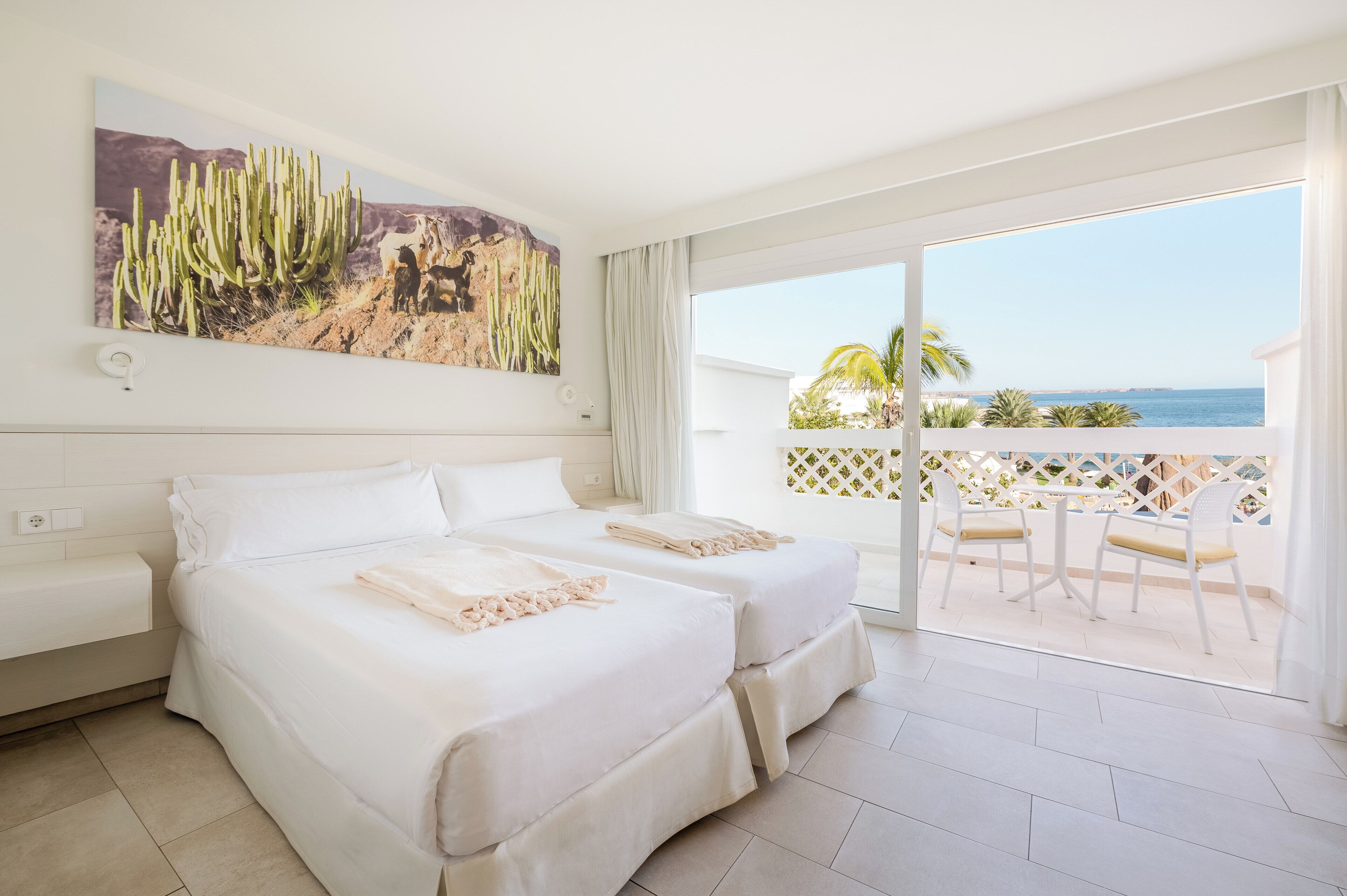 Ein Hotelzimmer mit zwei Betten, einem Balkon mit Stühlen und einem großen Wandbild einer Kaktee.