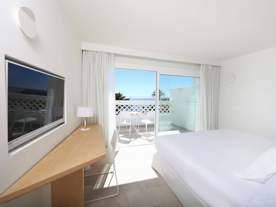 Habitación de hotel con cama, escritorio, silla, televisor y balcón con vista al mar.