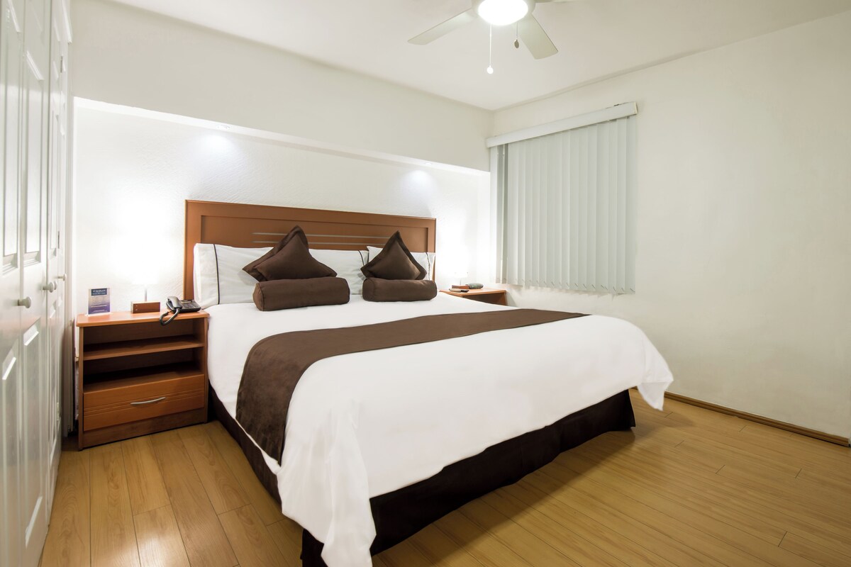 Una habitación de hotel con una cama grande, cabecera de madera, mesitas de noche, un teléfono y un ventilador de techo.