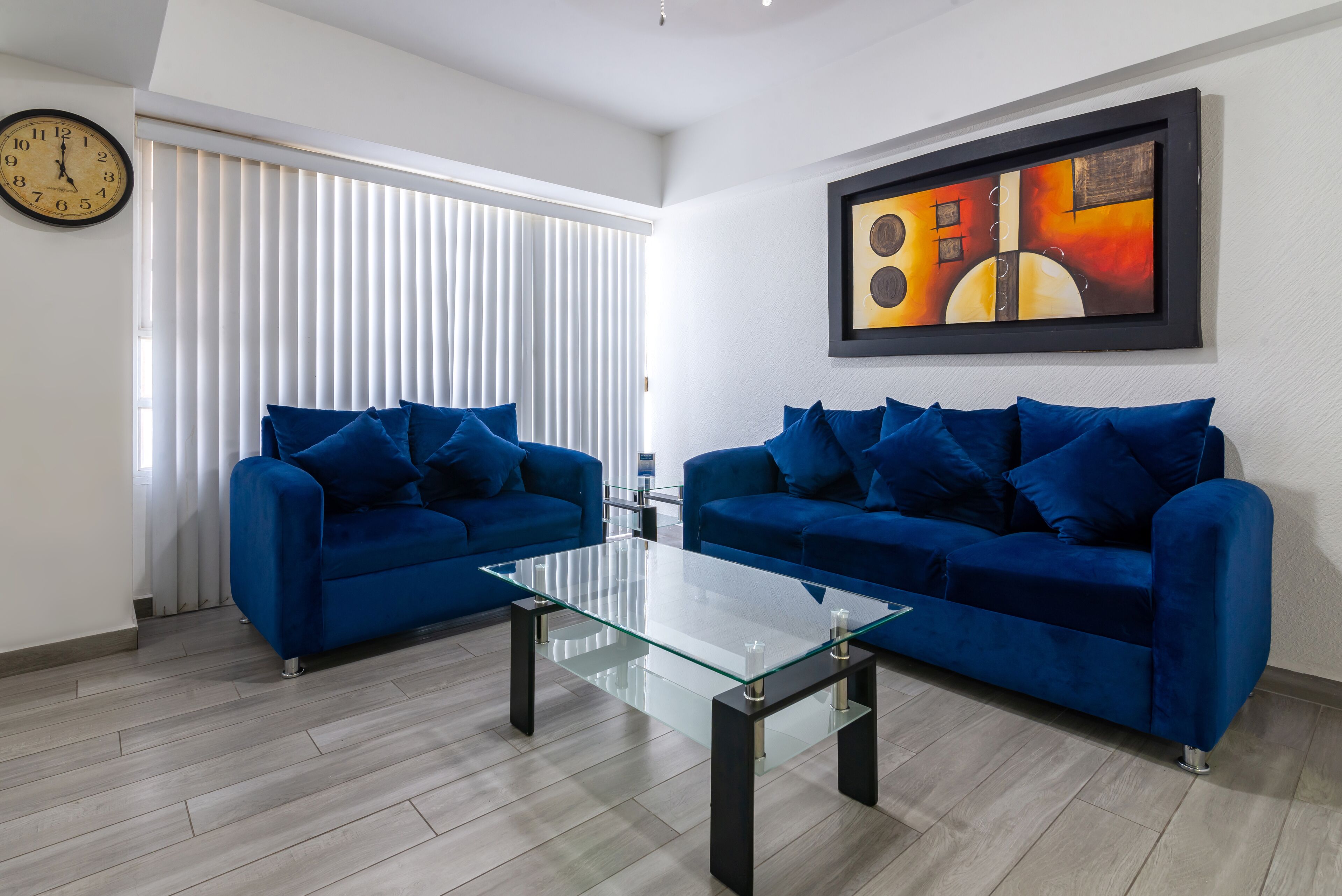 A modern living room with two blue sofas, a glass coffee table, a wall clock, and a framed abstract painting.