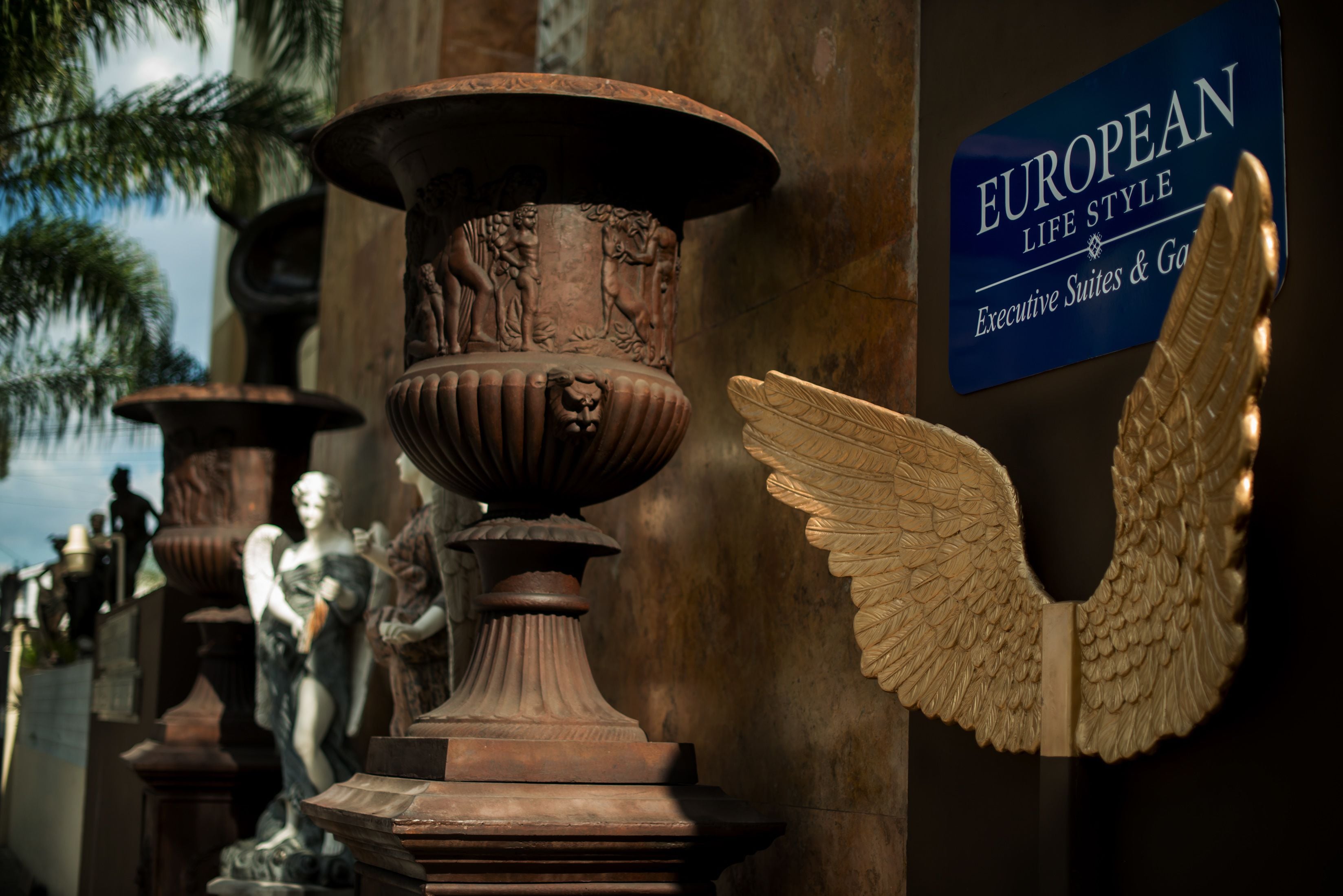 Une exposition de statues classiques et d’éléments architecturaux, comprenant une figure ailée et un panneau indiquant que le lieu est réservé aux suites et jardins de style européen, destinés à une clientèle aisée.