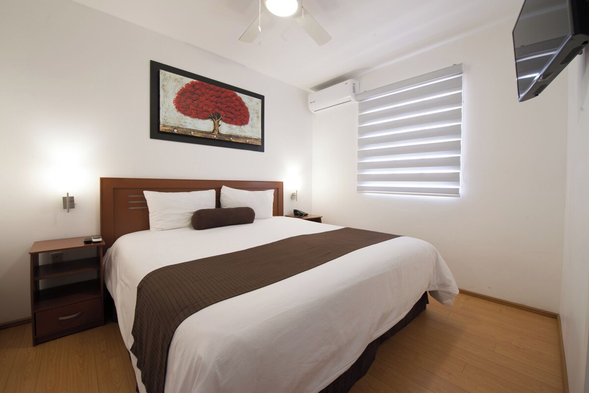 A hotel room with a bed, bedside tables, a nightstand, a wall-mounted TV, a ceiling fan, and a window with blinds.