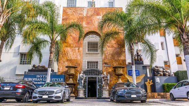 A hotel entrance with a large archway, palm trees, and parked cars. - European Life Style Executive Suites & Gallery (Zapopan)
