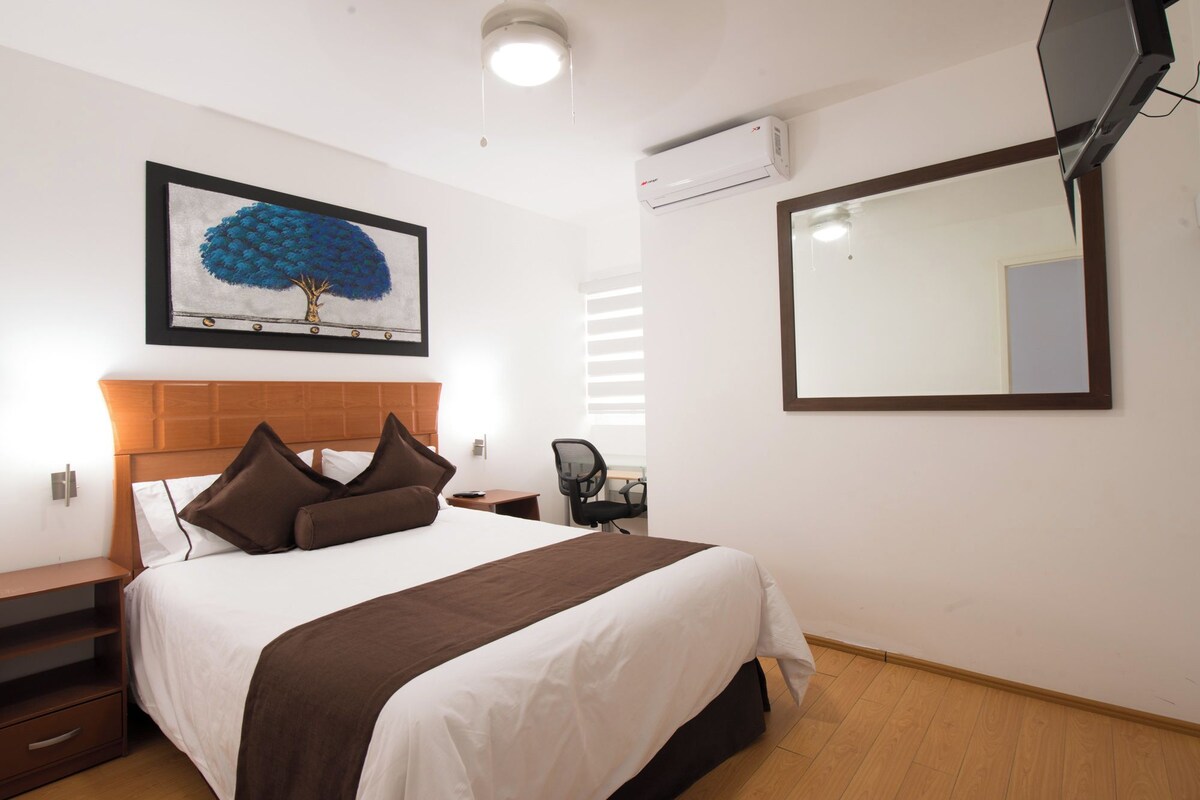 A hotel room with a bed, bedside table, chair, and a painting on the wall.