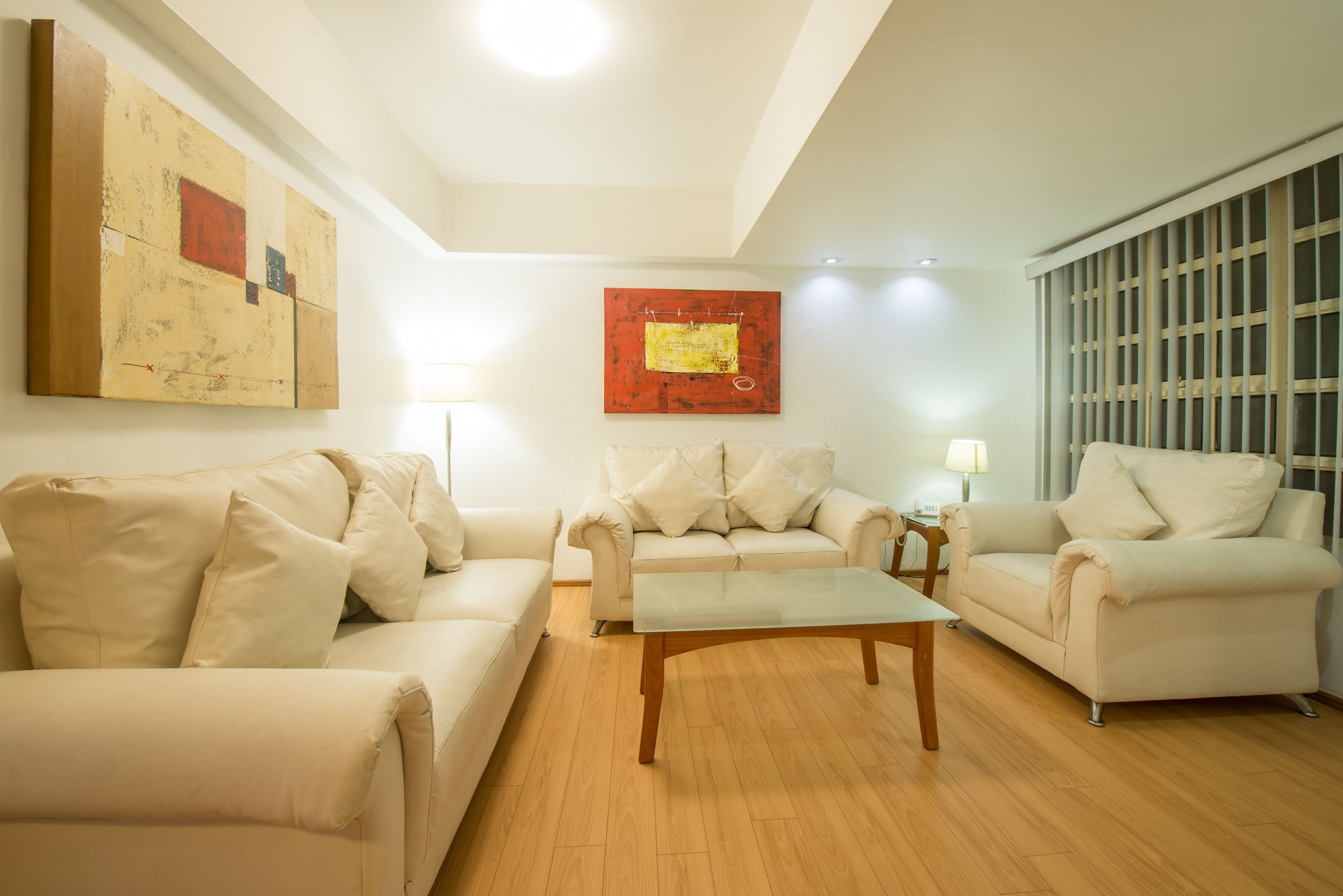 A living room with three beige sofas, a glass coffee table, and two wall-mounted lamps.