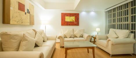 A living room with three beige sofas, a glass coffee table, and two wall-mounted lamps.